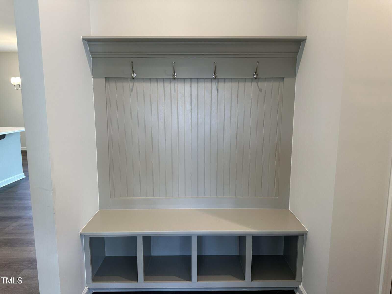 A built-in mudroom bench with four open cubbies underneath, a paneled back, and a shelf above featuring four metal hooks for hanging items, set against a neutral wall.