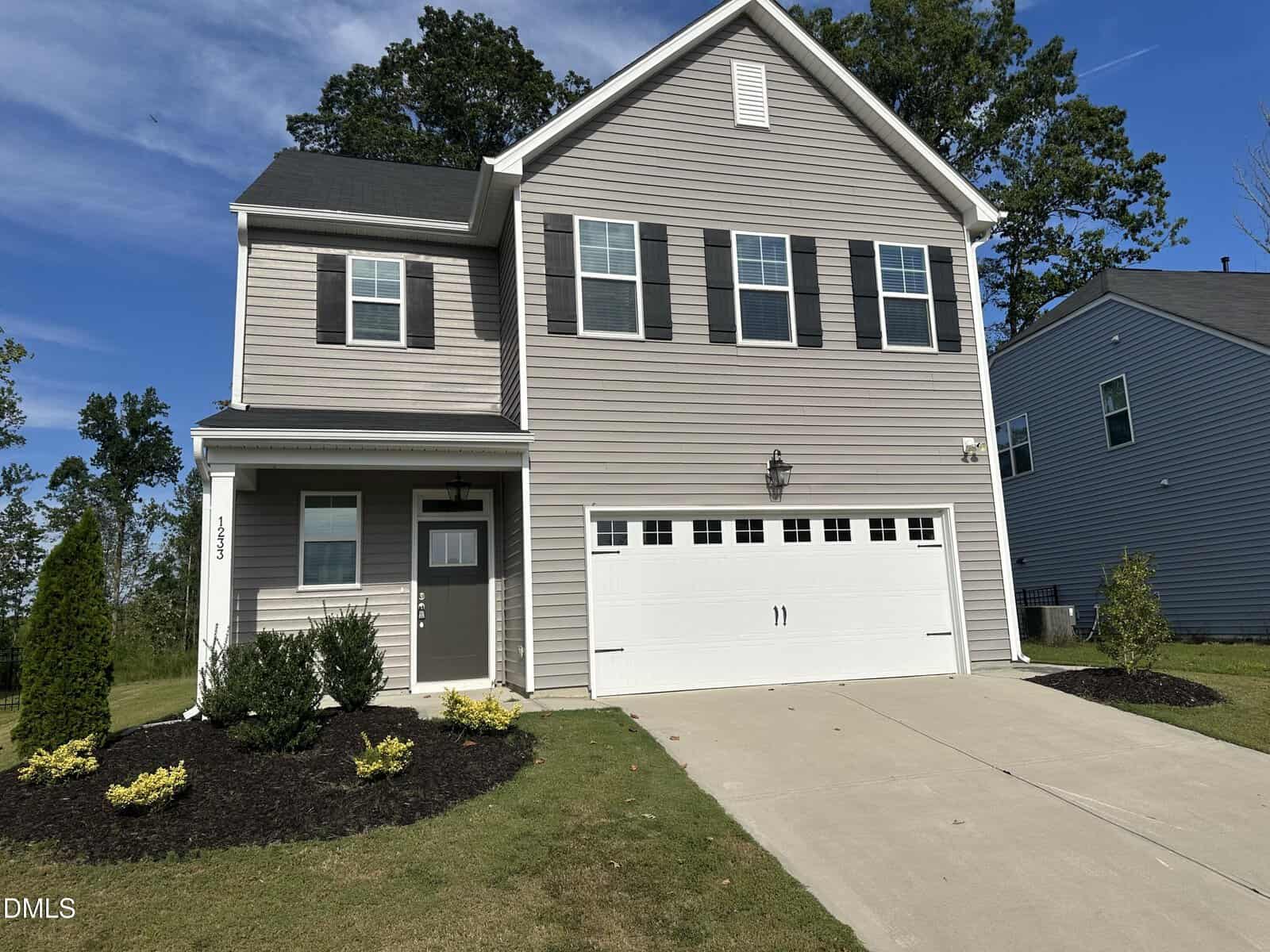 Nestled in Wake Forest, this two-story beige house at 1233 Shadow Shade Drive features black shutters, white trim, a double garage, and sits on a manicured lawn with shrubs and fresh mulch on a sunny day.