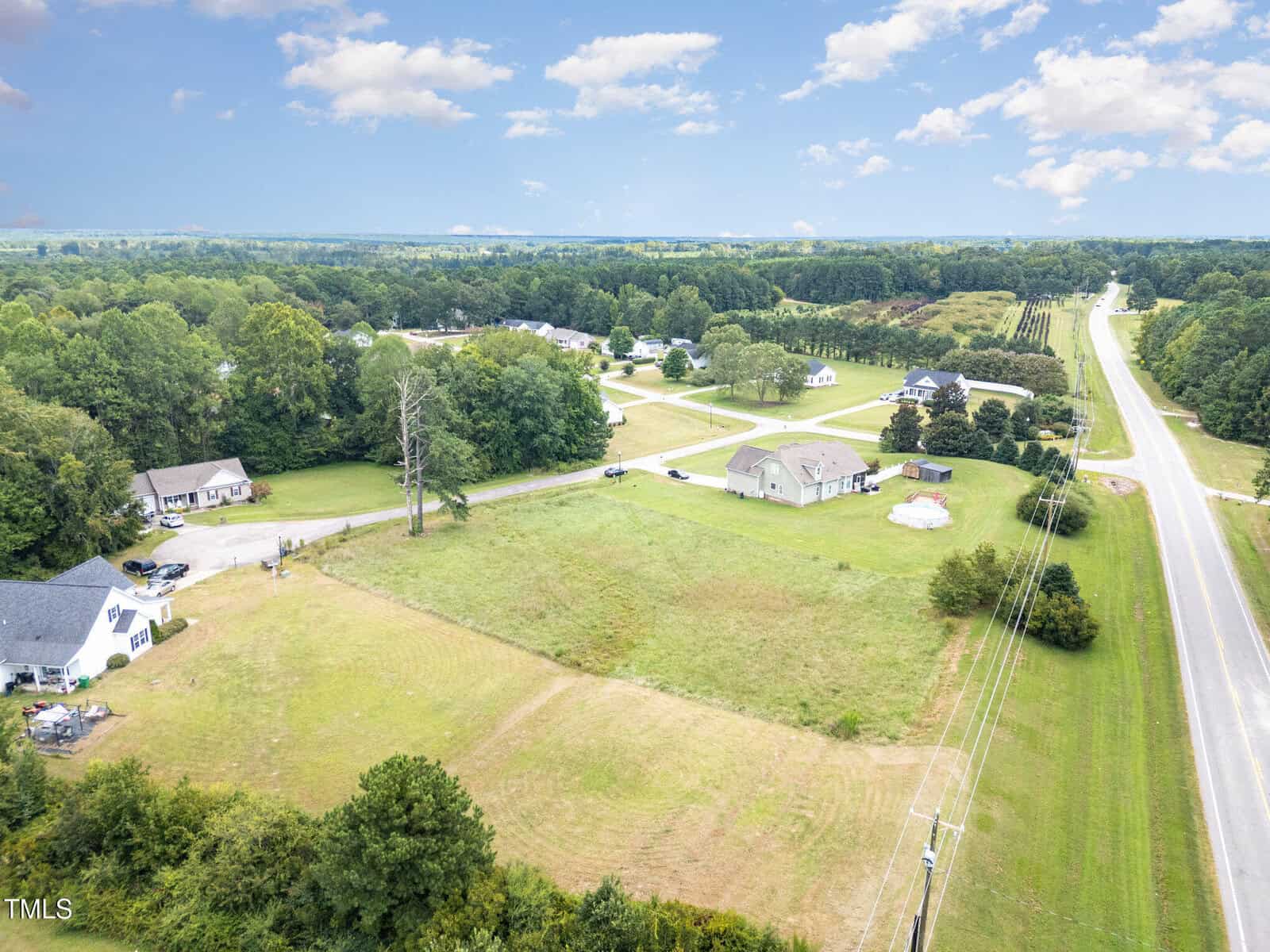 Aerial view of a rural neighborhood with several houses, open grassy fields, scattered trees, and a main road running along the right side of the image under a partly cloudy sky.