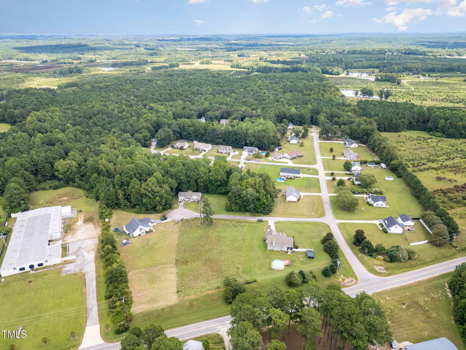 Aerial view of a rural residential neighborhood with houses, green lawns, trees, and intersecting roads. Open fields and a large industrial building are visible along the edges of the community.