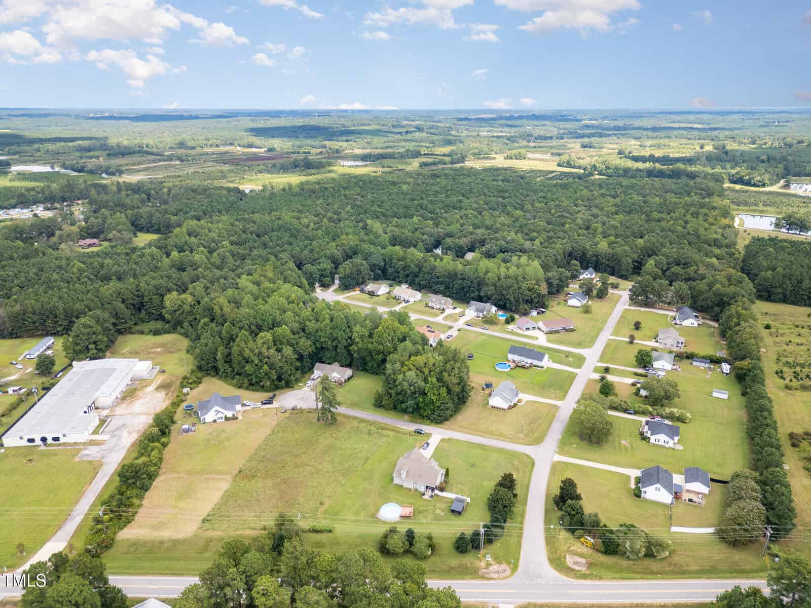 Aerial view of a rural residential neighborhood with scattered houses, open fields, and wooded areas, intersected by paved roads under a partly cloudy sky.