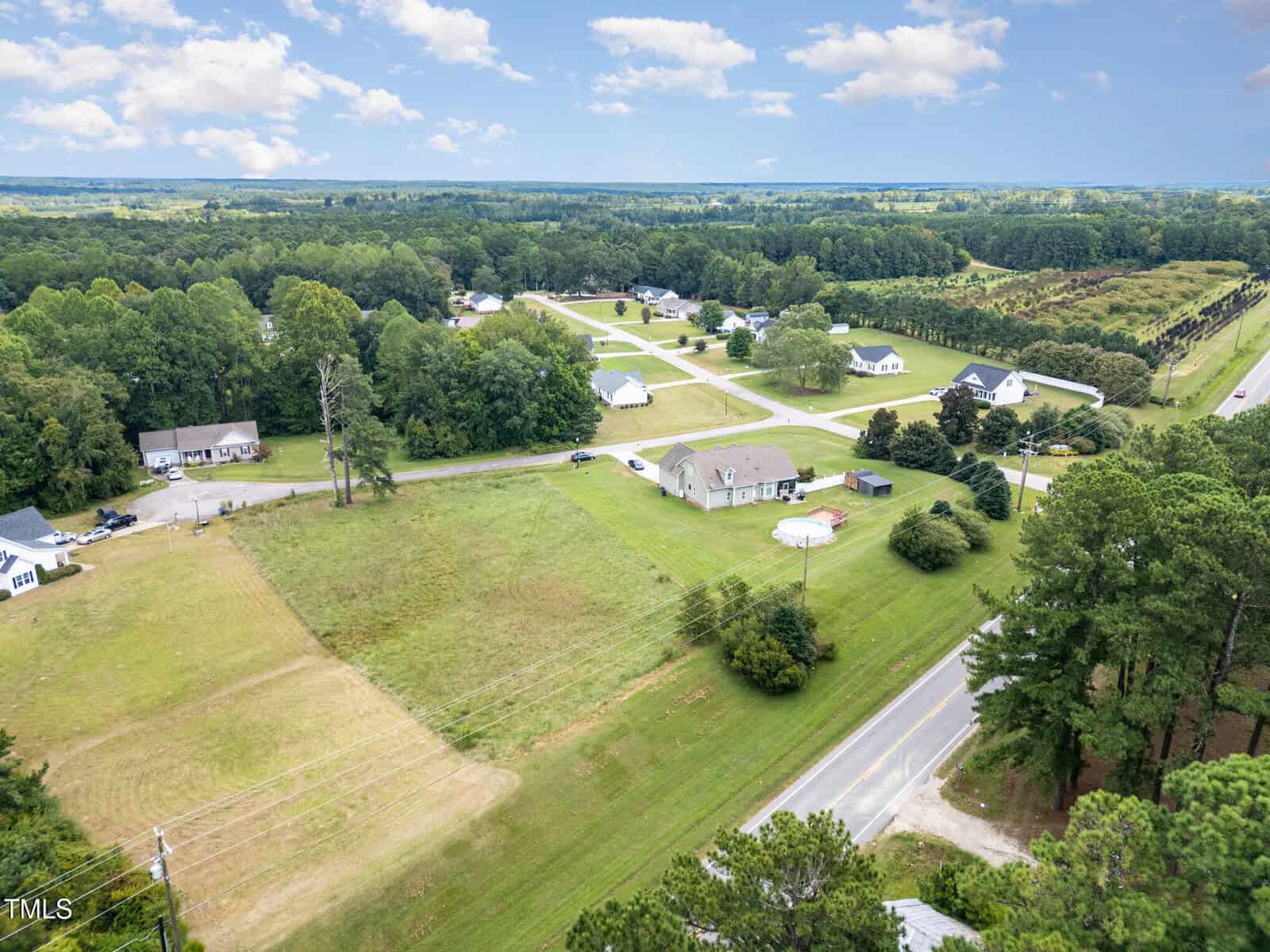 Aerial view of a rural neighborhood with several houses, open grassy fields, a few trees, and intersecting roads under a partly cloudy sky.