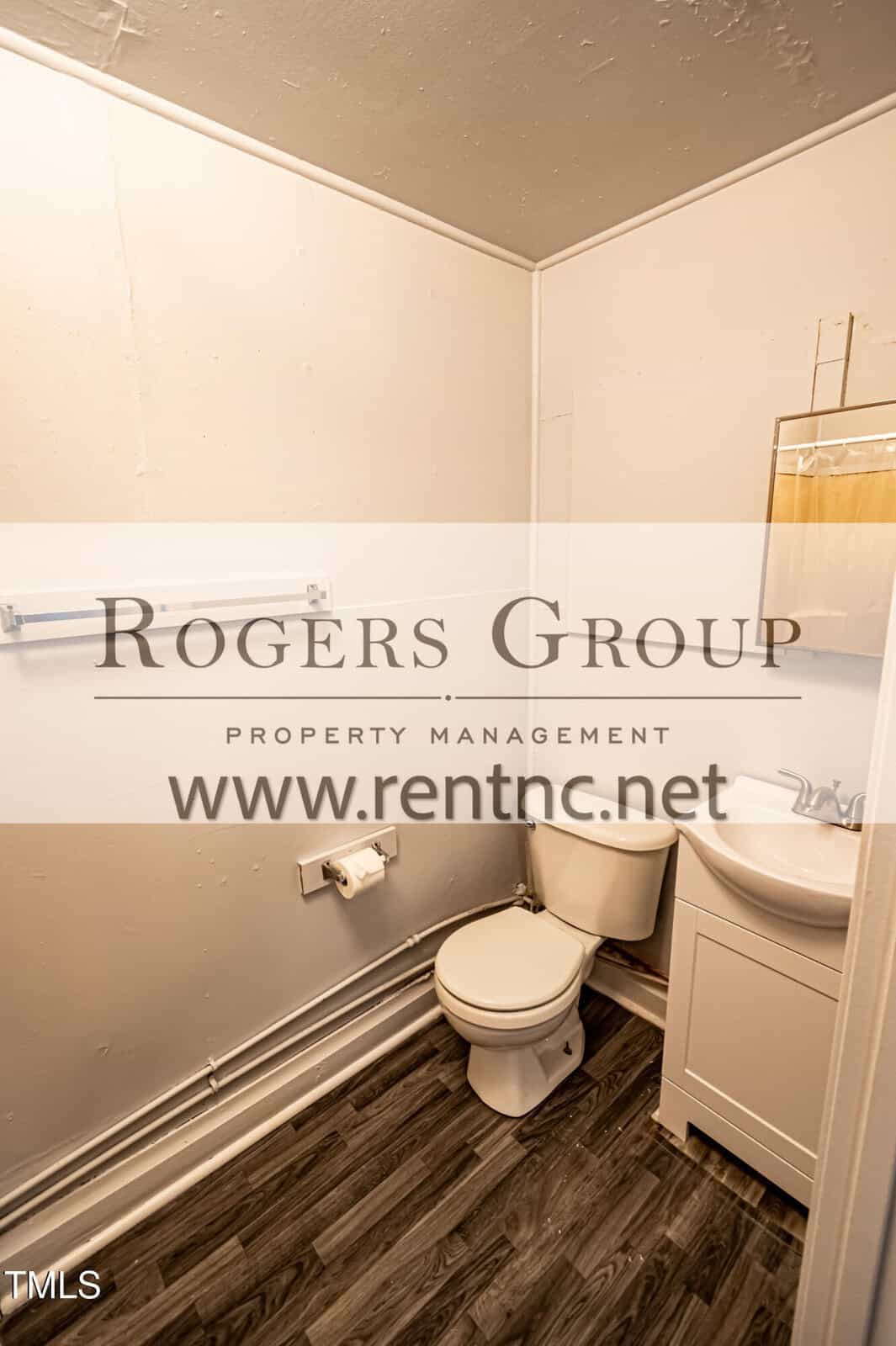 A small bathroom with a white toilet, a white sink cabinet, a towel bar, and a toilet paper holder, featuring light-colored walls and dark wood-style flooring. The Rogers Group logo and website are overlaid on the image.