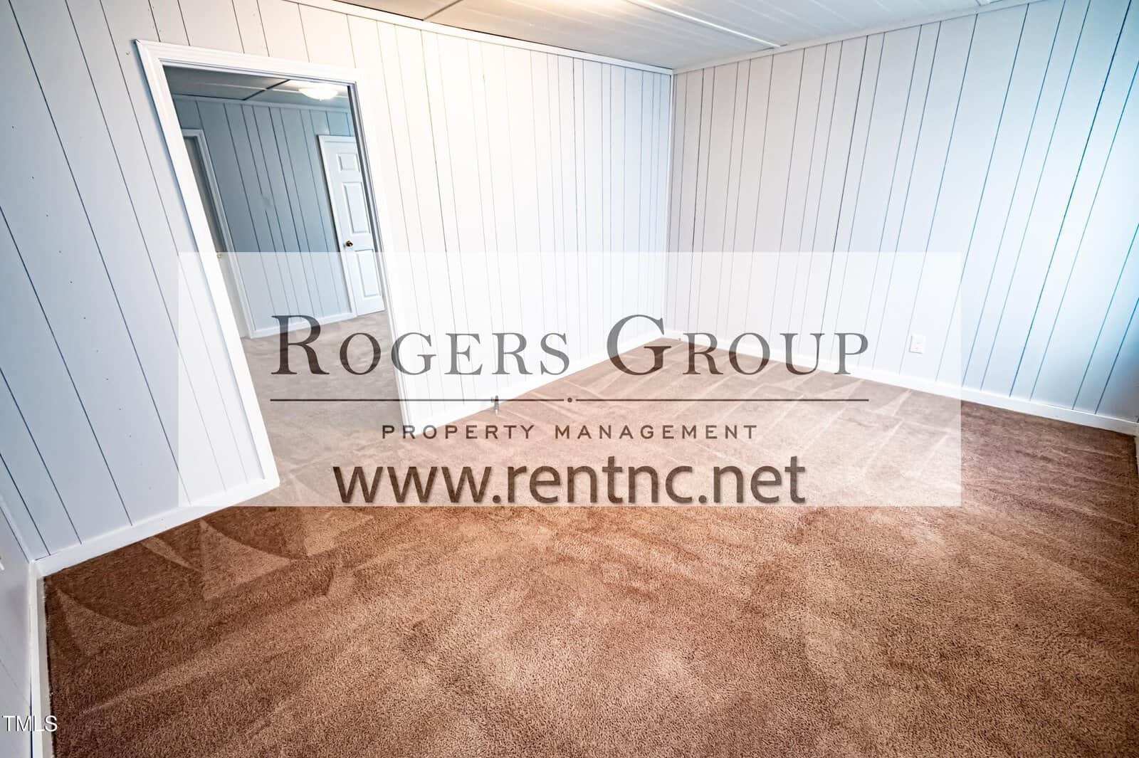 Empty room with beige carpet, white paneled walls, and a white ceiling. There is an open doorway leading to another room. A logo for Rogers Group Property Management and a website overlay the image.