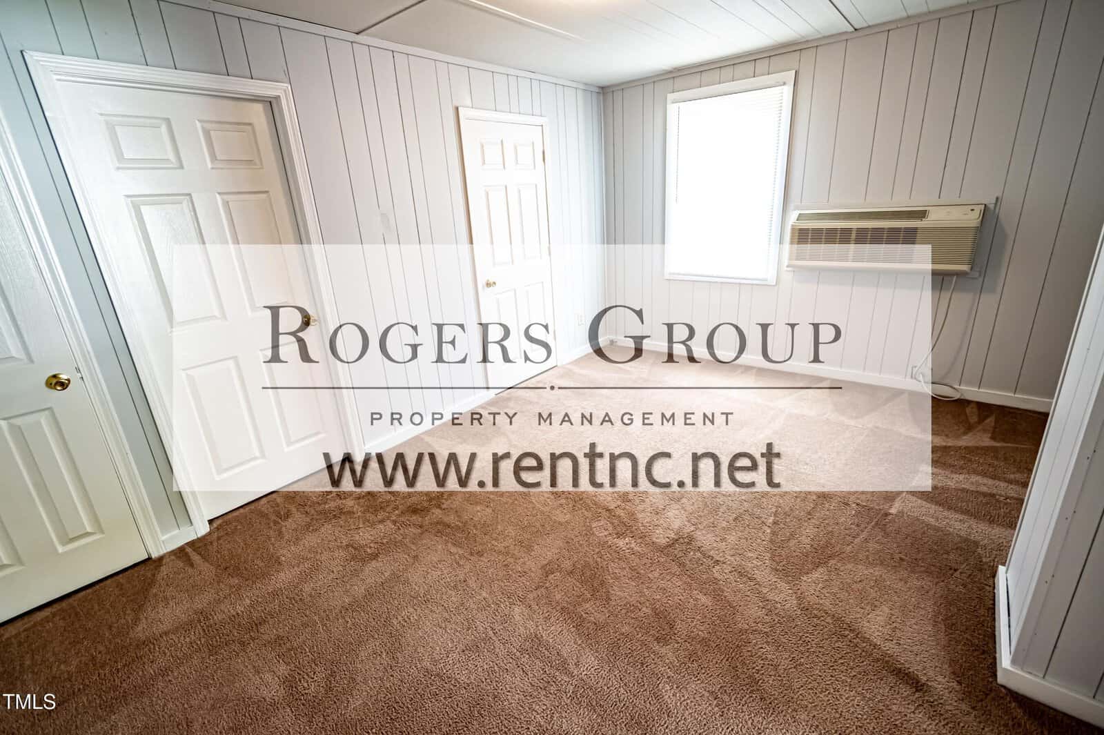 A small, empty room with carpeted floors, light gray wood-paneled walls, two white doors, a window, and a wall-mounted air conditioning unit. The Rogers Group logo and website are overlaid on the image.