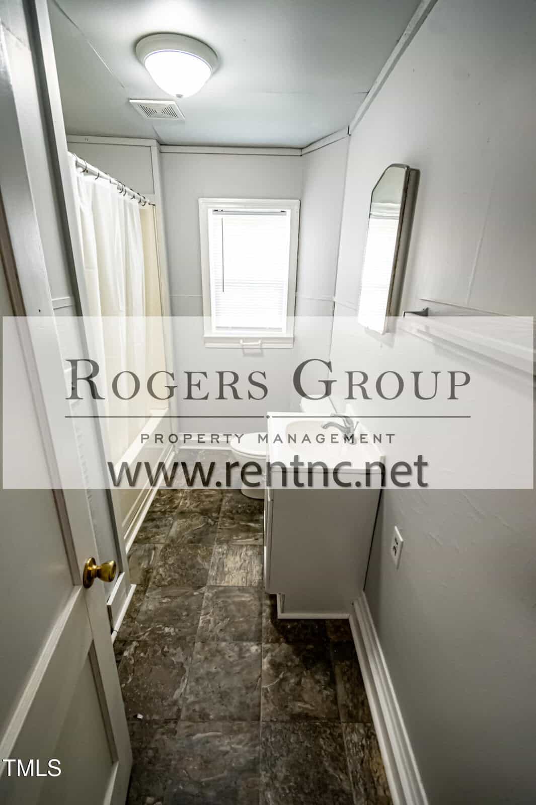 A narrow bathroom with gray tile flooring, a small white vanity with a mirror above it, a window with blinds, and a shower curtain on the left. The Rogers Group property management watermark is over the image.