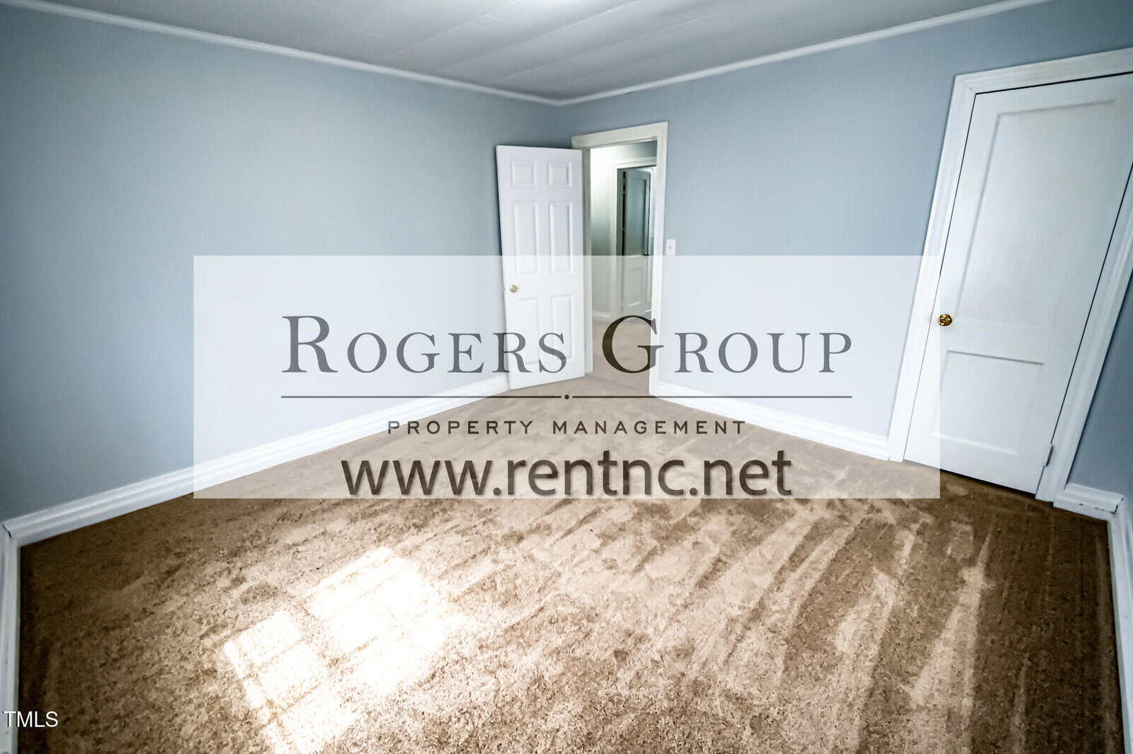 Empty room with light blue walls, beige carpet, white trim, an open door, and a closed closet door. Rogers Group Property Management ad overlay with website www.rentnc.net.