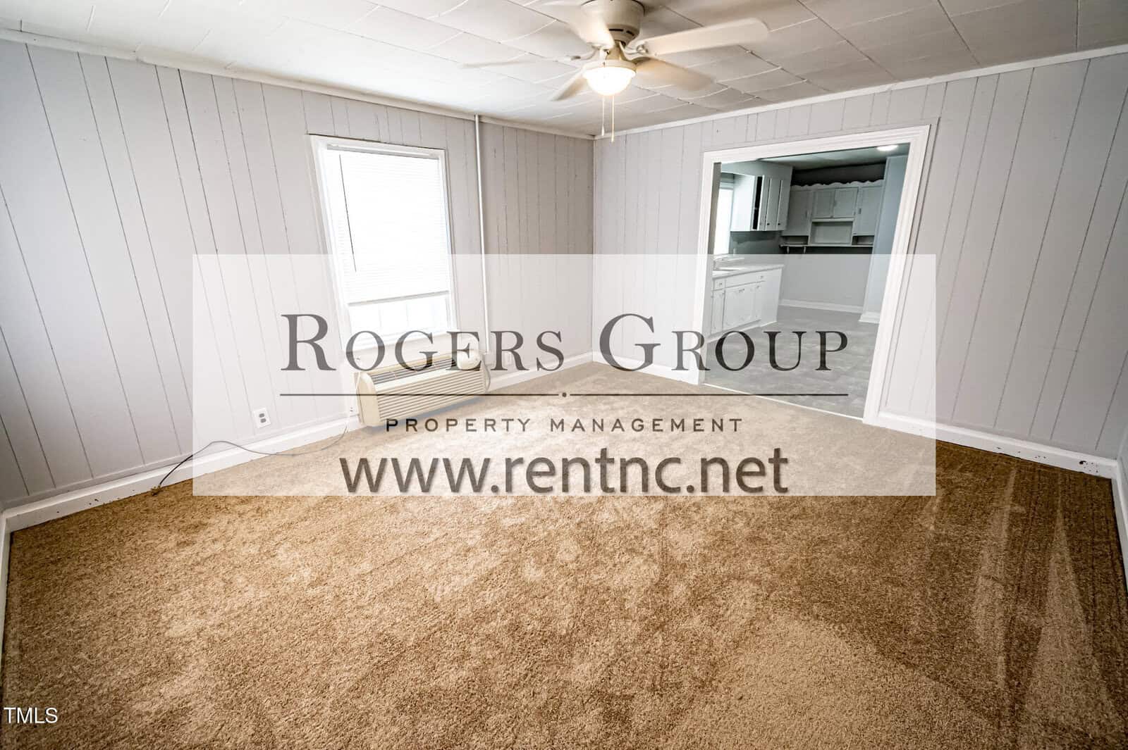 Empty room with tan carpet, light gray paneled walls, two windows, a ceiling fan, and an open doorway leading to a kitchen. Rogers Group Property Management logo and website appear over the image.