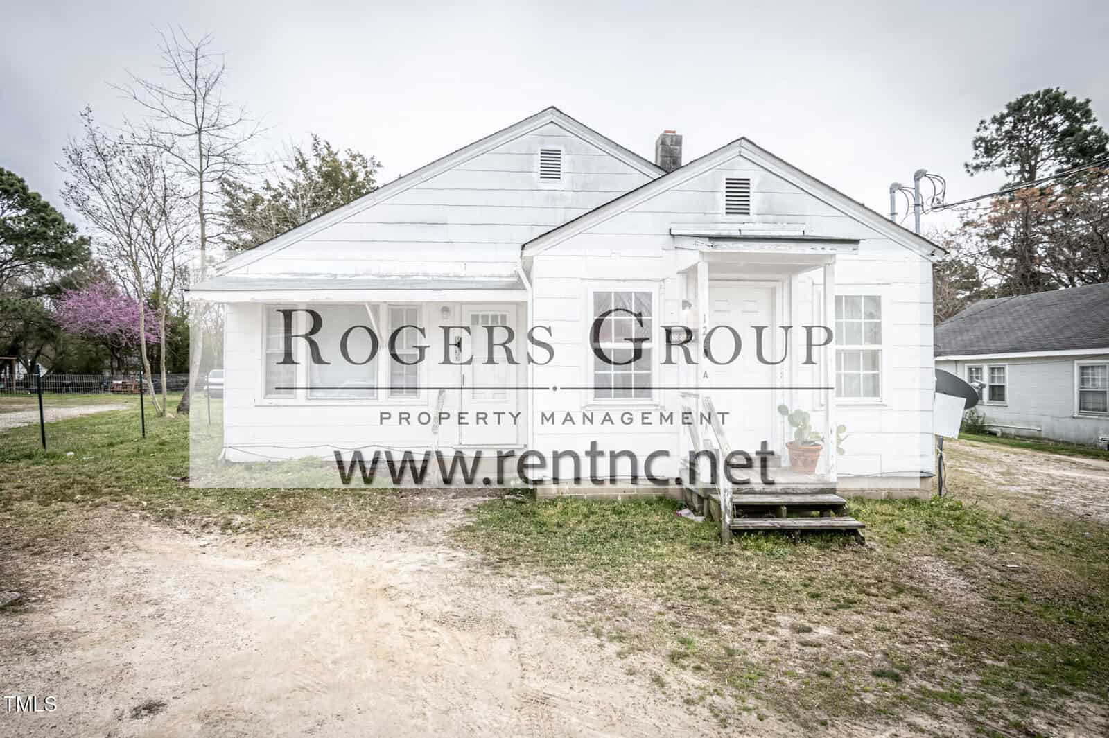 A small, white, single-story house with a front porch and two front doors, situated on a patchy grass and dirt yard. The house has simple exterior features and a few potted plants near the steps.