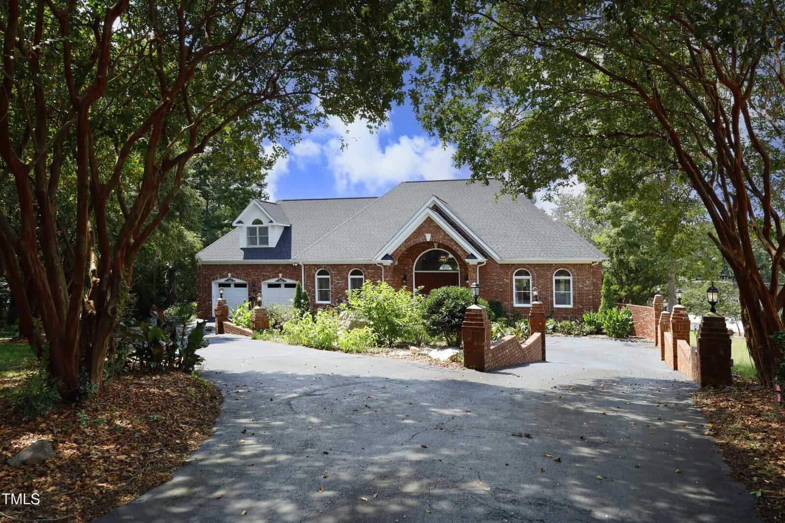 A large brick house with arched windows and a covered entryway sits at the end of a curved driveway, framed by tall trees and lush greenery under a bright blue sky.