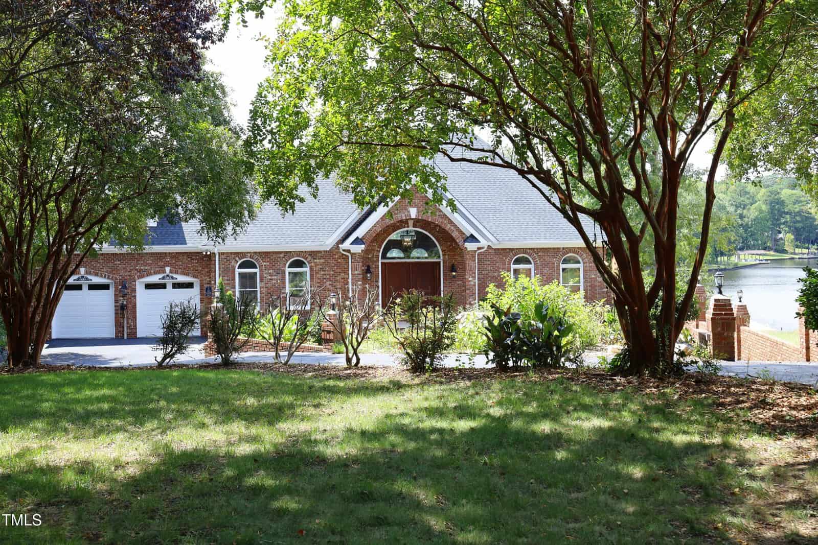 A brick house with arched windows and a double garage is partially shaded by trees in the front yard. The house sits near a body of water, visible on the right side of the image.
