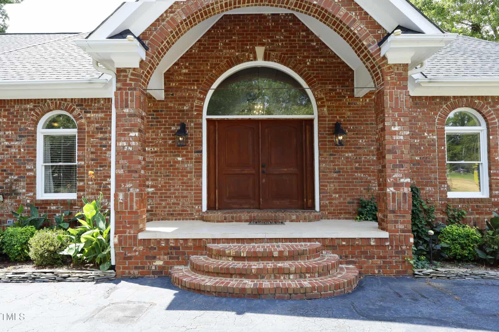 A red brick house entrance with double wooden doors, arched transom window above, two black wall lanterns, and curved brick steps leading up to the front porch, surrounded by green plants.