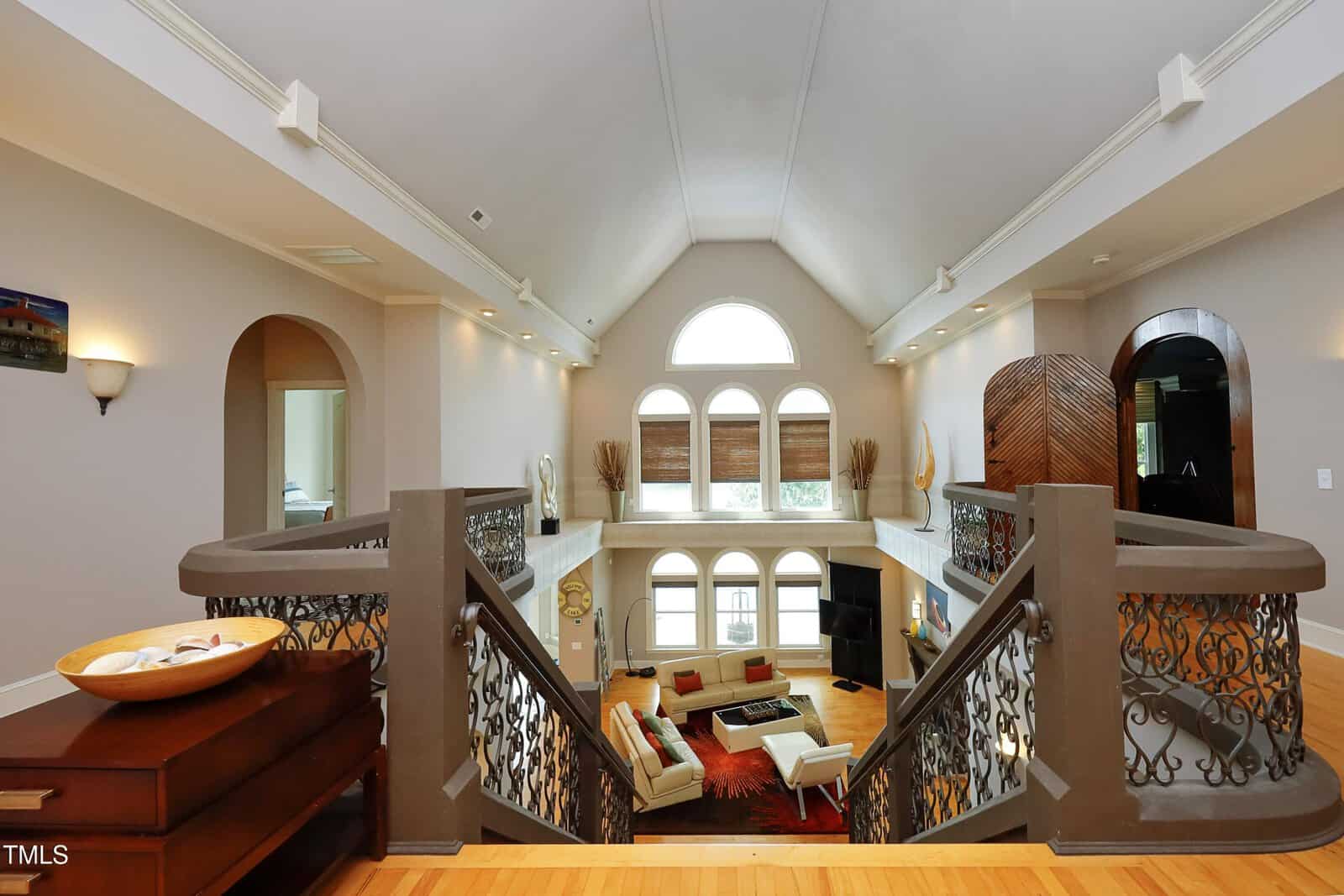 View from the top of a staircase overlooking a spacious living room with vaulted ceilings, large arched windows, modern furniture, and decorative railings on both sides of the stairs.