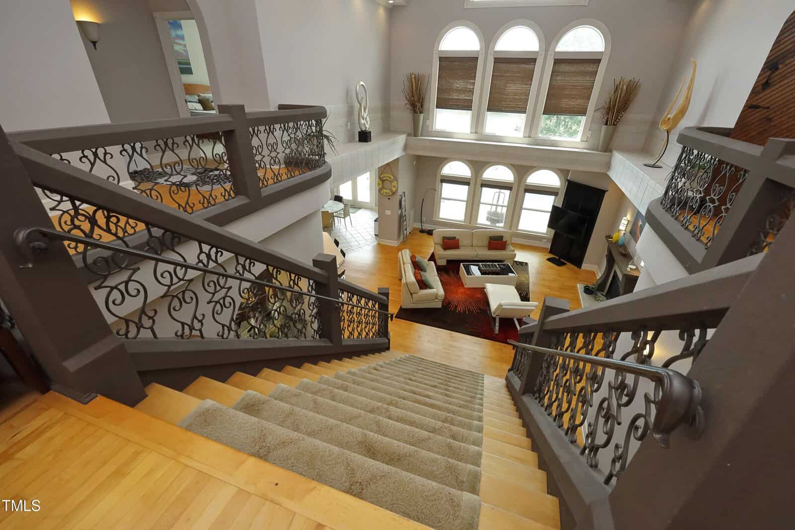 View from the top of a carpeted staircase in a spacious, contemporary living room with high ceilings, large arched windows, modern decor, a sectional sofa, and ornate iron railings.