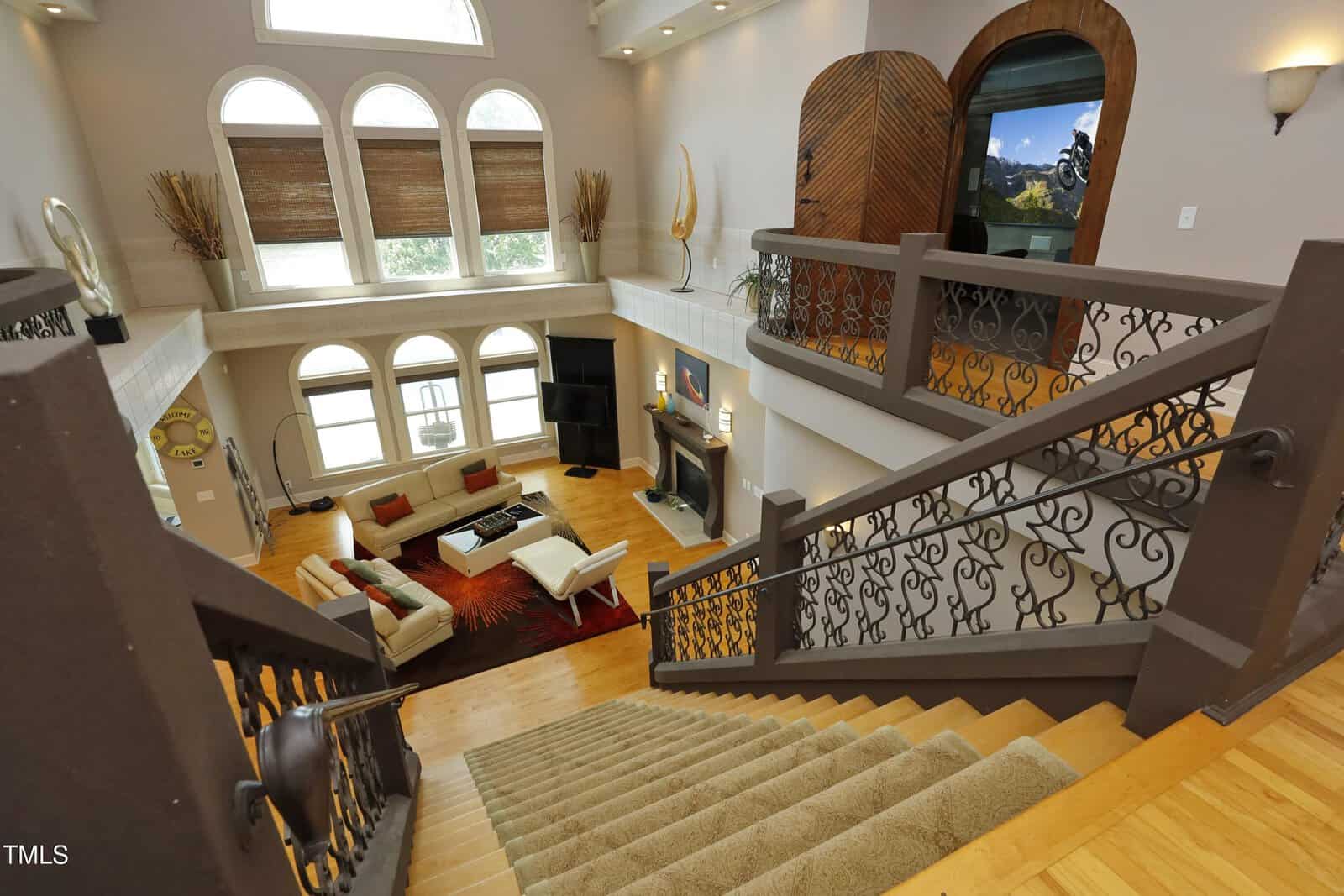 View from a staircase overlooking a spacious, modern living room with high ceilings, large windows, contemporary furniture, and decorative accents, including a fireplace and wall art.