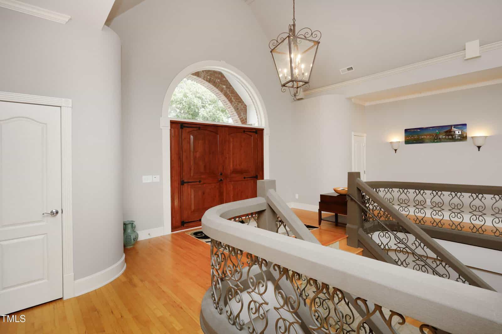 Grand foyer with a large wooden double door, ornate staircase railing, hardwood floors, high ceiling, chandelier, wall sconces, and a landscape photo hanging on a light gray wall.