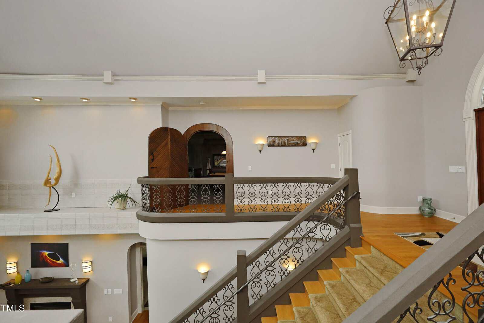 Second-floor hallway overlooks staircase with ornate railing. Modern art, sculpture, and decorative lighting accent the beige and gray walls. Hardwood floors contrast with tan carpet on the stairs. A large lantern hangs above.