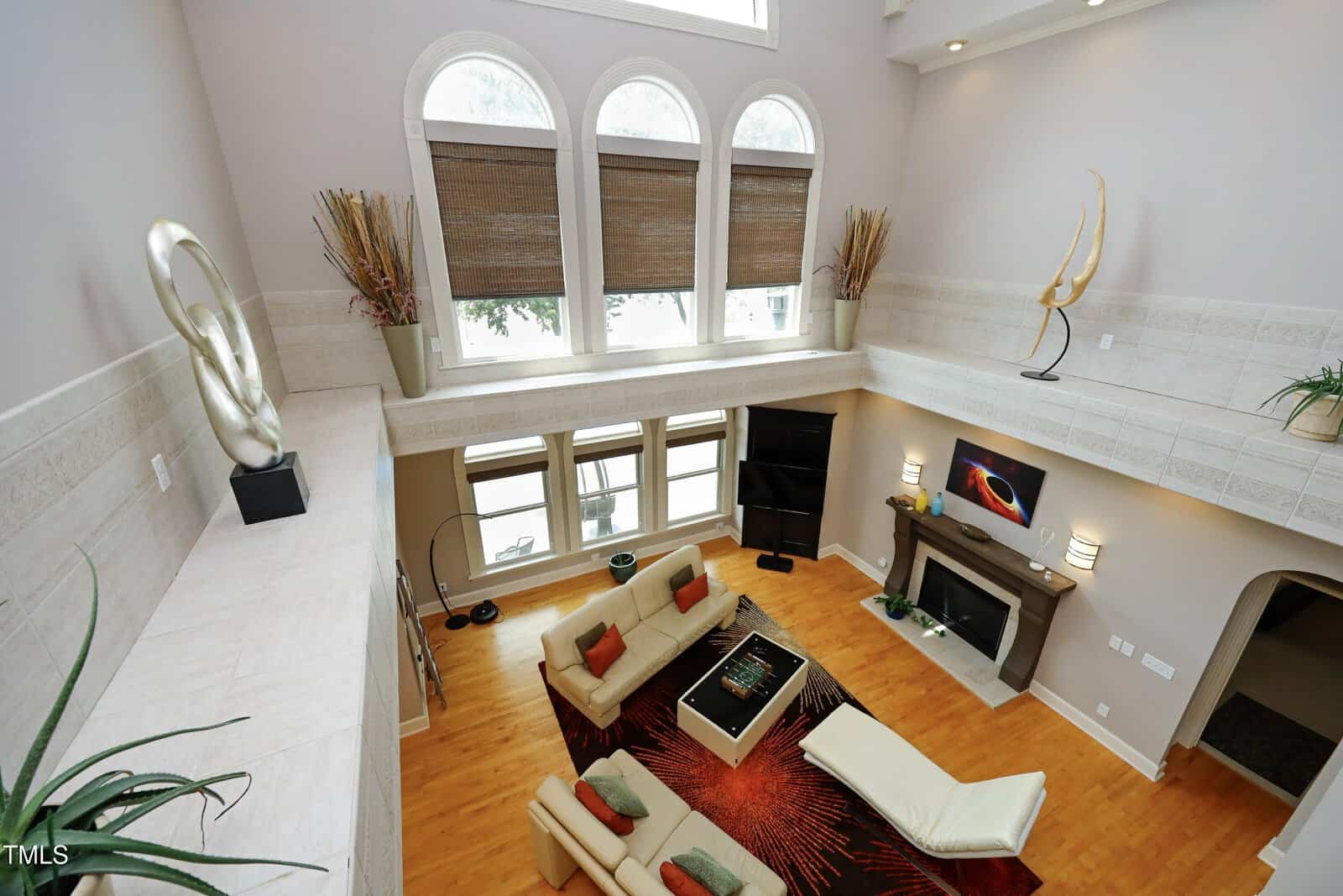 Modern living room with tall windows, a fireplace, contemporary art, cream seating, and orange-accented decor. Warm wooden floor and light walls create an open, airy atmosphere.