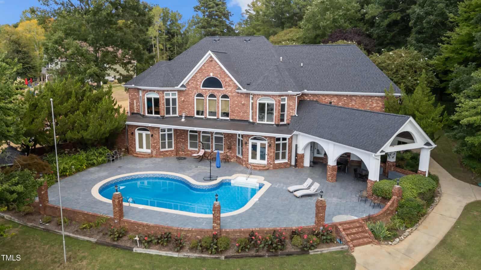 A large two-story brick house at 120 Chuckwagon Drive in Louisburg overlooks a backyard with a kidney-shaped swimming pool, gray stone patio, lounge chairs, and lush greenery and trees.