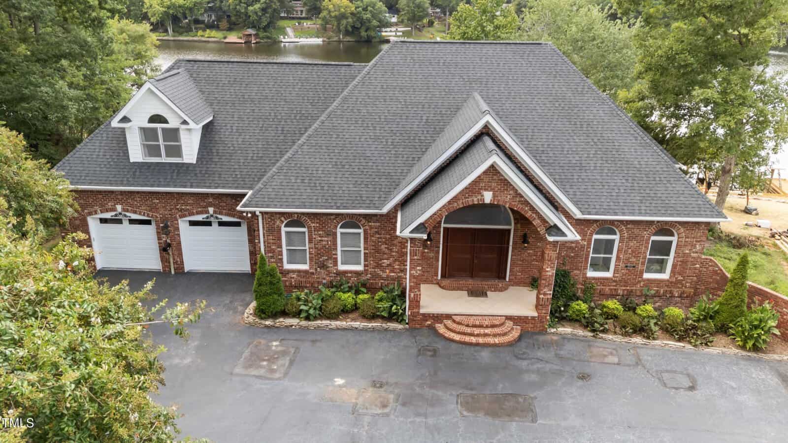 A large brick house with a dark gray roof, double garage, arched windows, and a covered front entry sits in front of a lake, surrounded by trees and a paved driveway.