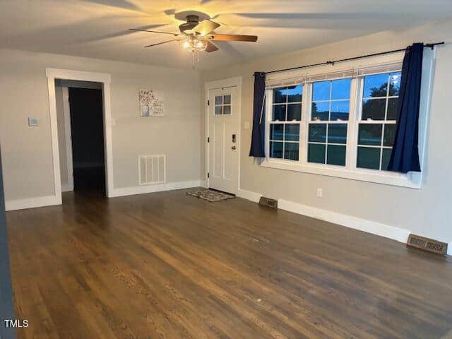Empty living room with wooden floors, white walls, large window with blue curtains, ceiling fan with lights, and a white front door. A small mat is by the door, and wall art hangs next to an interior door.