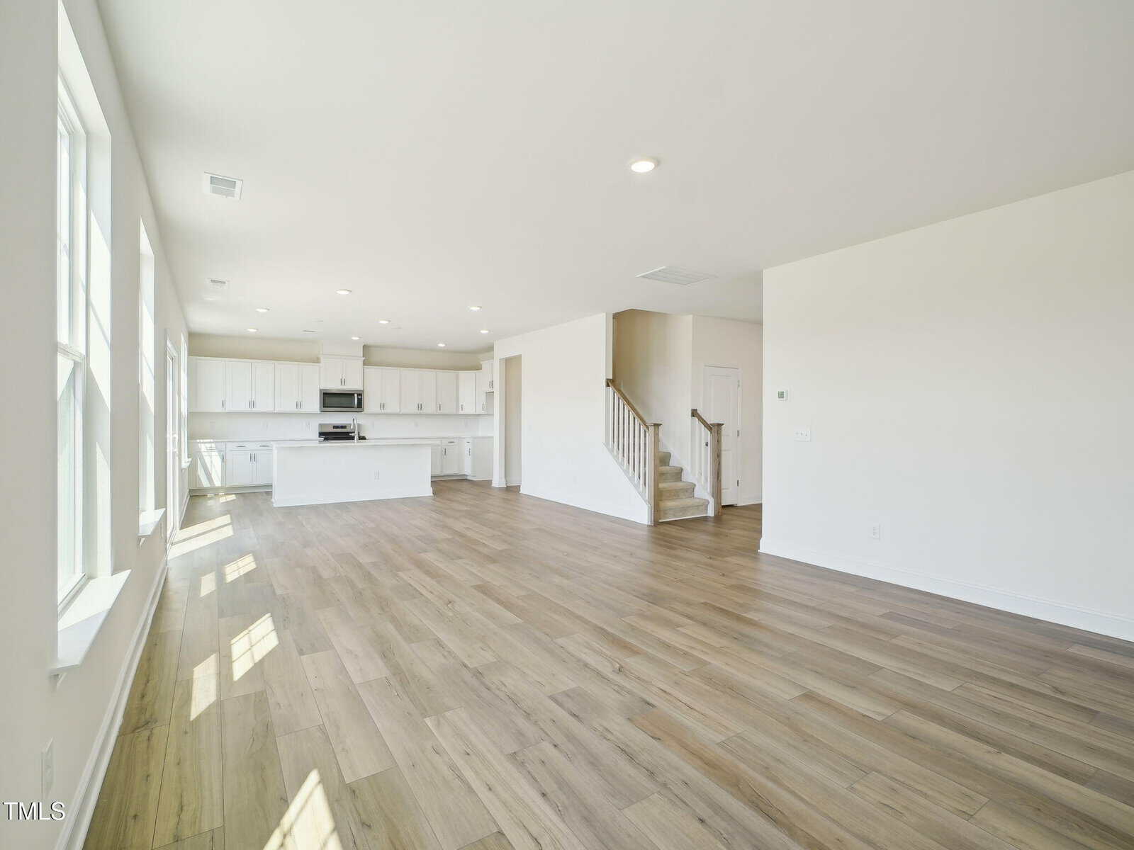 Spacious, empty open-concept living area with light wood floors, white walls, large windows, and a modern kitchen with white cabinets and a center island. Carpeted staircase leads to an upper floor.