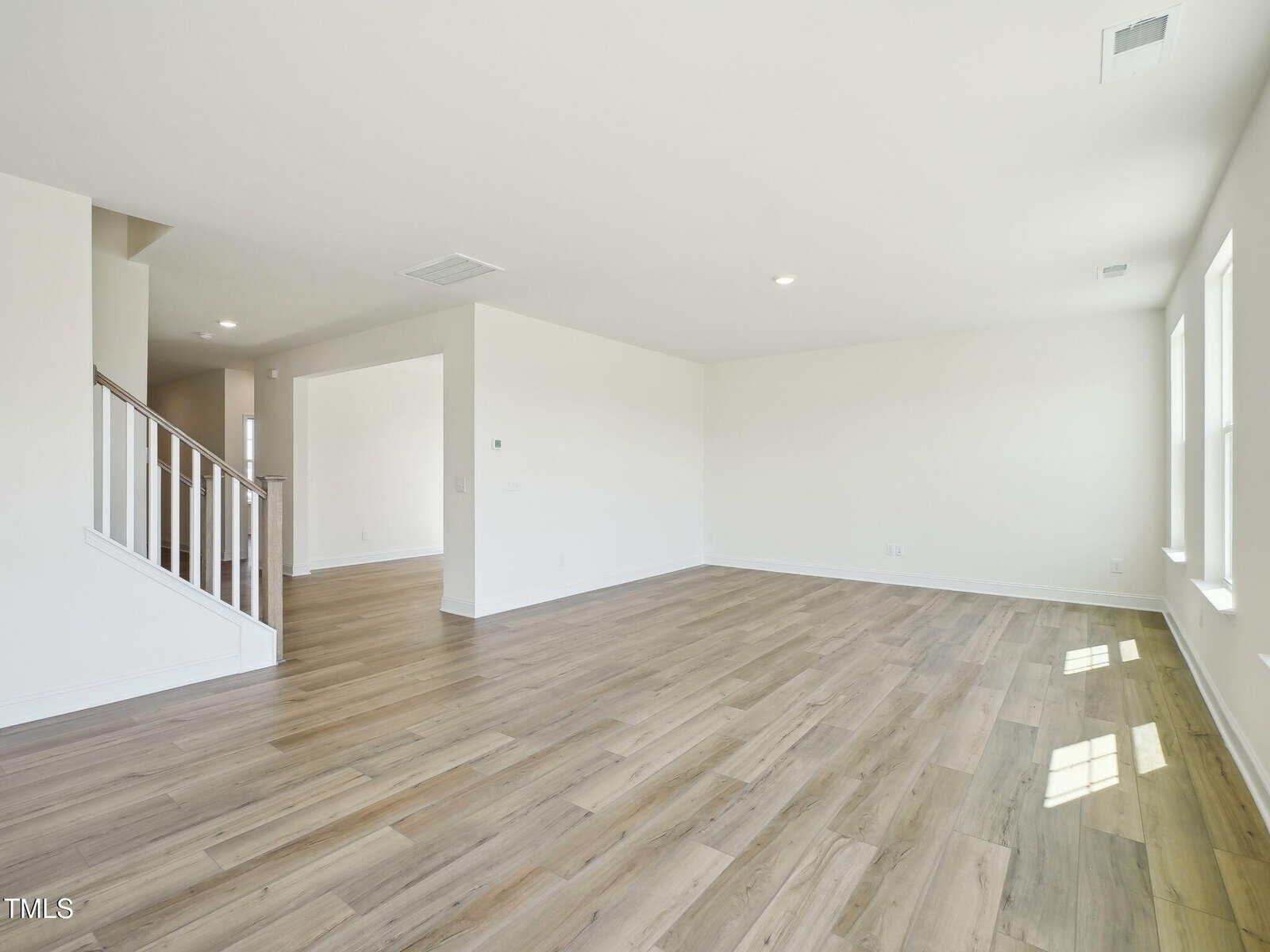 Empty, unfurnished living room with light wood flooring, off-white walls, recessed lighting, a staircase with white railing, two windows letting in natural light, and an open doorway leading to another room.