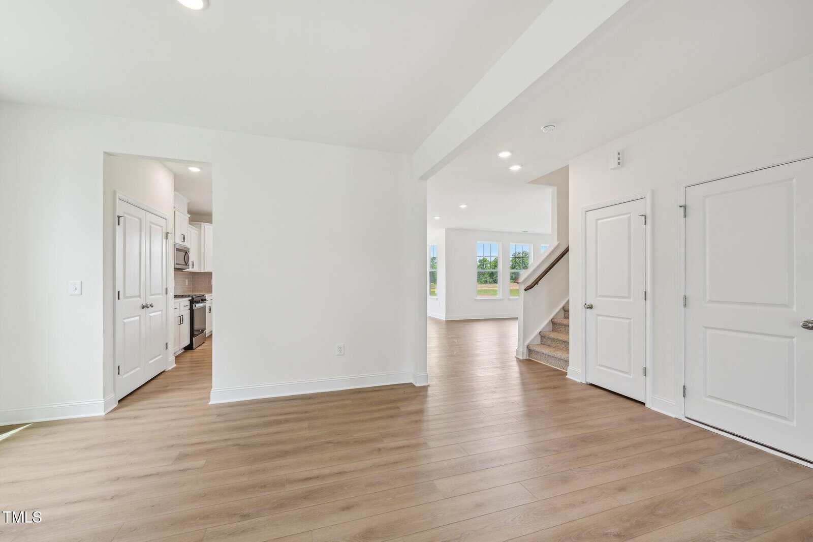 Bright, modern interior with light wood floors, white walls, a kitchen with stainless steel appliances to the left, carpeted stairs leading up, and a living area with large windows in the background.