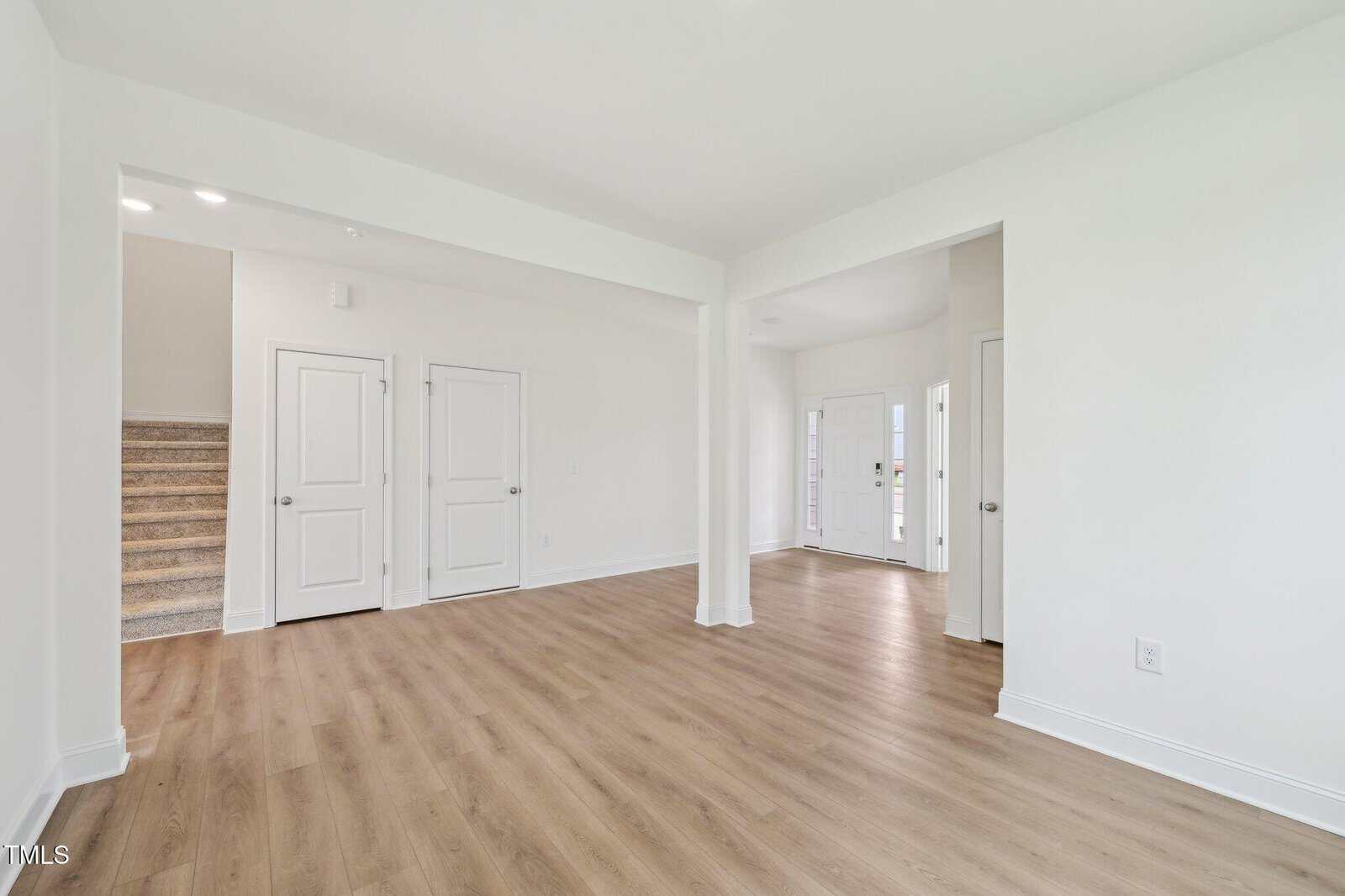 A bright, empty room with white walls, light wood flooring, two closed doors, a staircase with carpeted steps, and an open entryway leading to a front door with sidelights.