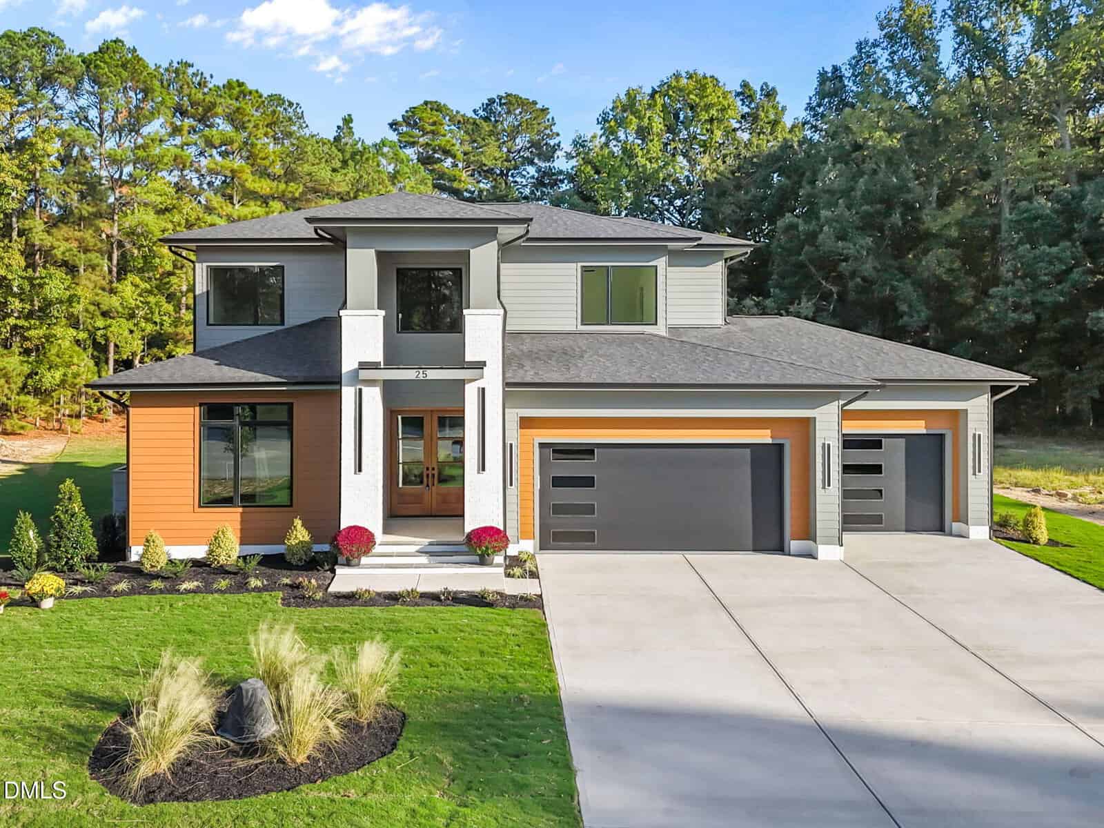 A modern two-story house at 25 Geranium Drive, Youngsville, with a gray and wood exterior, three-car garage, large windows, and landscaped front yard, set against tall trees and a clear sky.