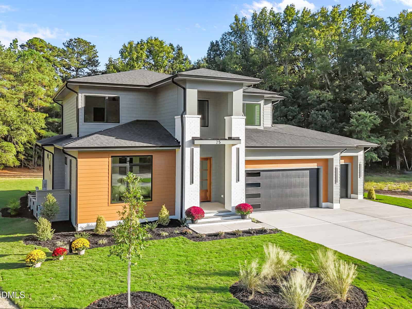 A modern two-story house with gray and wood-paneled exterior, large windows, and a balcony. The front yard is landscaped with grass, plants, and shrubs, and there is a spacious driveway and a three-car garage.