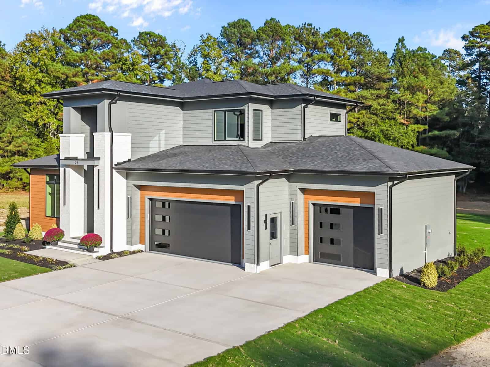 Modern two-story house with gray siding, large black-framed windows, and three black garage doors. The home has a wide driveway, minimal landscaping, and is surrounded by tall trees in the background.