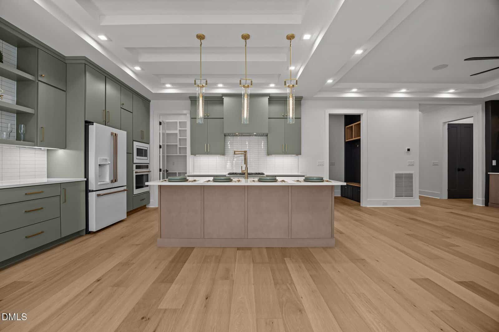 Modern kitchen with sage green cabinets, a large island with a beige base, white countertops, gold hardware, three pendant lights above the island, white appliances, wood flooring, and recessed ceiling lights.