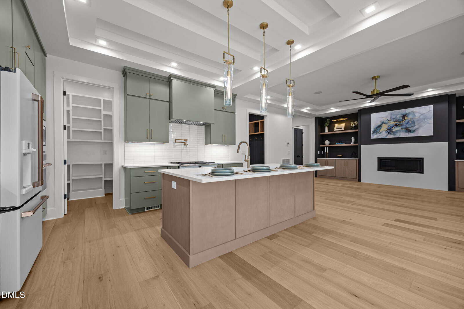 Modern kitchen with a large island, green cabinets, open pantry, pendant lights, built-in shelves, fireplace, ceiling fan, and wood flooring. Contemporary decor with neutral tones.