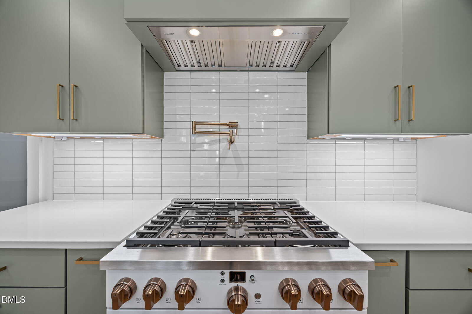 Modern kitchen with a stainless steel gas stove, brass knobs, white countertops, olive green cabinets, white subway tile backsplash, and a brass pot filler faucet above the stove.