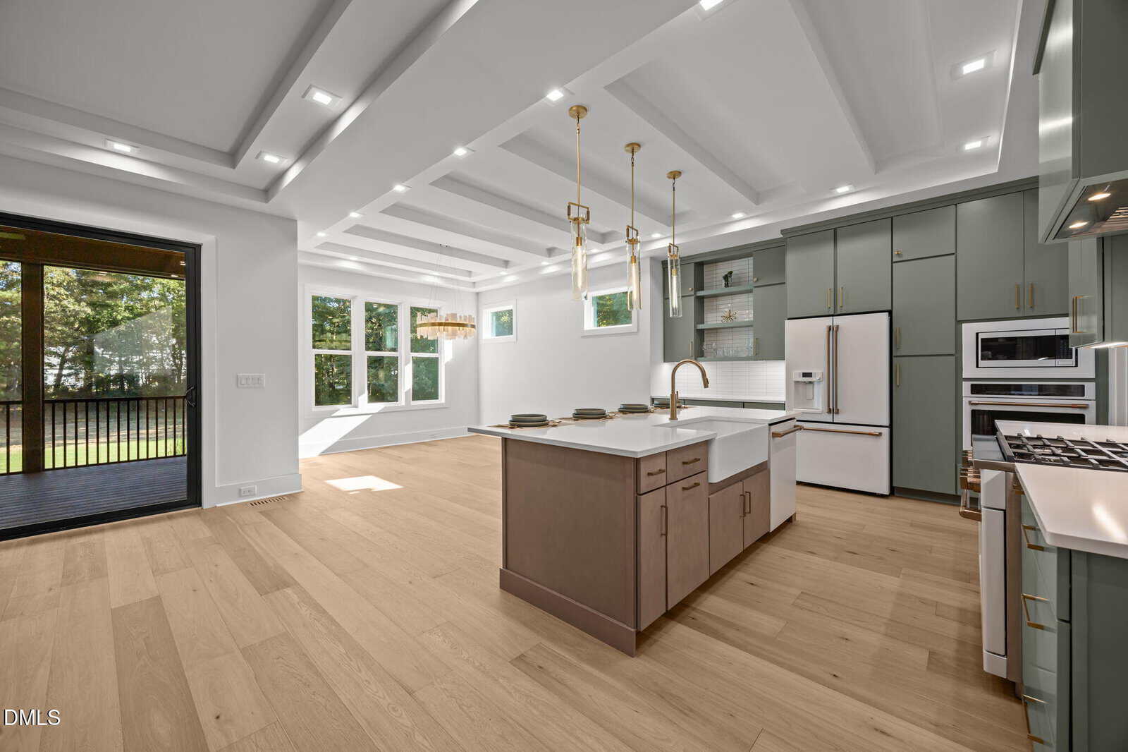 Modern kitchen with light wood floors, green cabinetry, a large island with a sink, gold pendant lights, white countertops, built-in appliances, and large windows overlooking a grassy outdoor area.