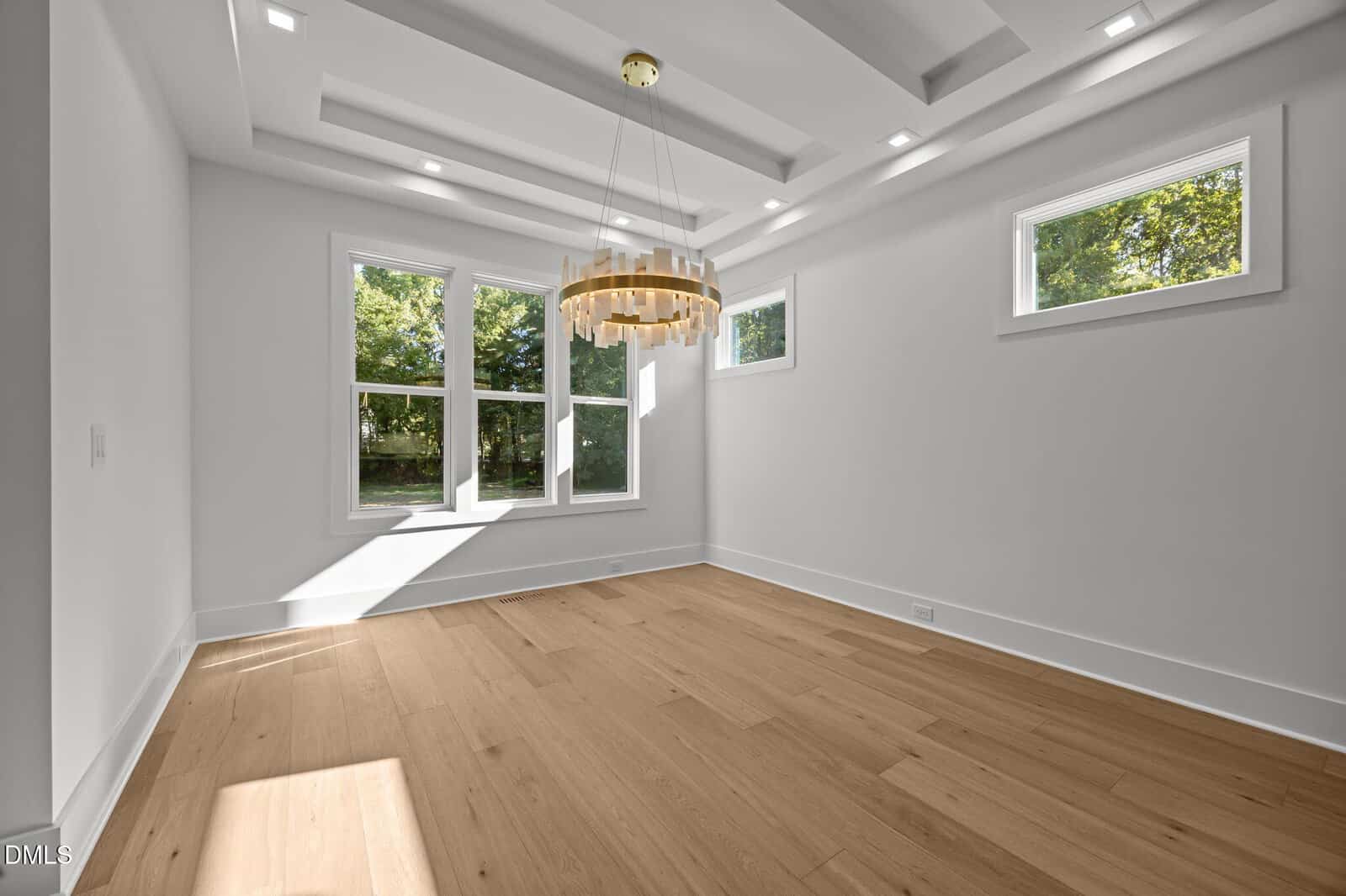 Bright, modern empty room with light wood floors, white walls, large windows letting in sunlight, square recessed ceiling lights, and a stylish circular chandelier hanging from a coffered ceiling.