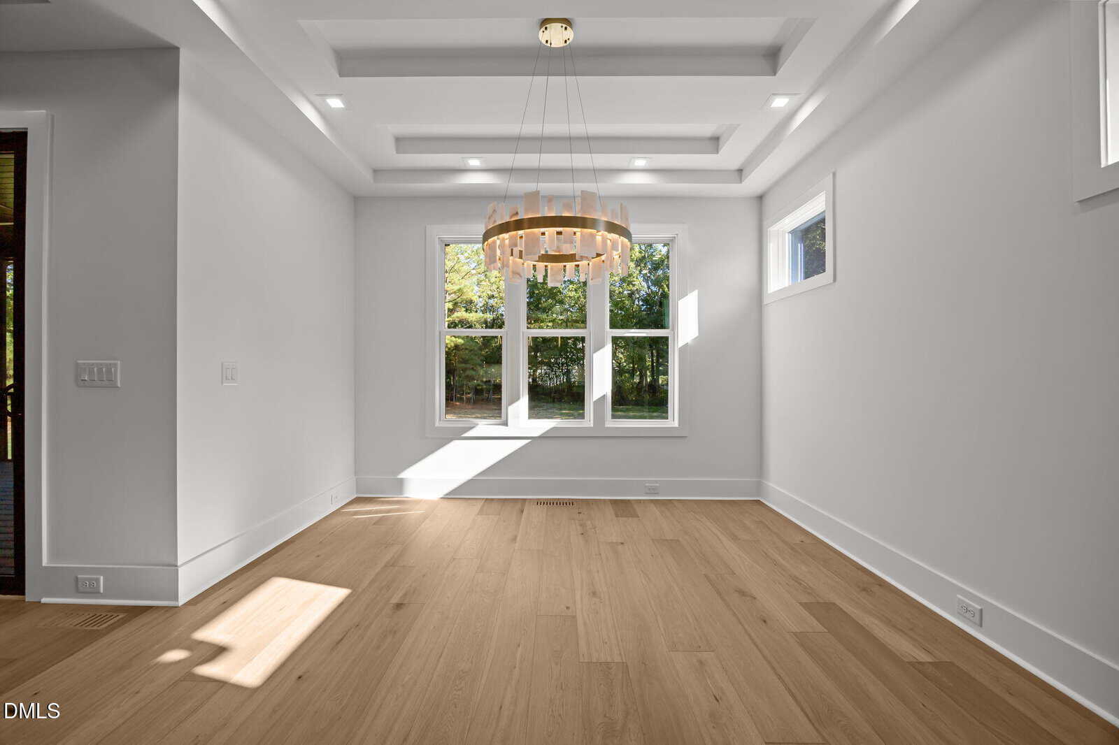A bright, empty room with light wood floors, white walls, large windows, ceiling tray details, and a circular modern chandelier. Sunlight streams through the windows, casting shadows on the floor.