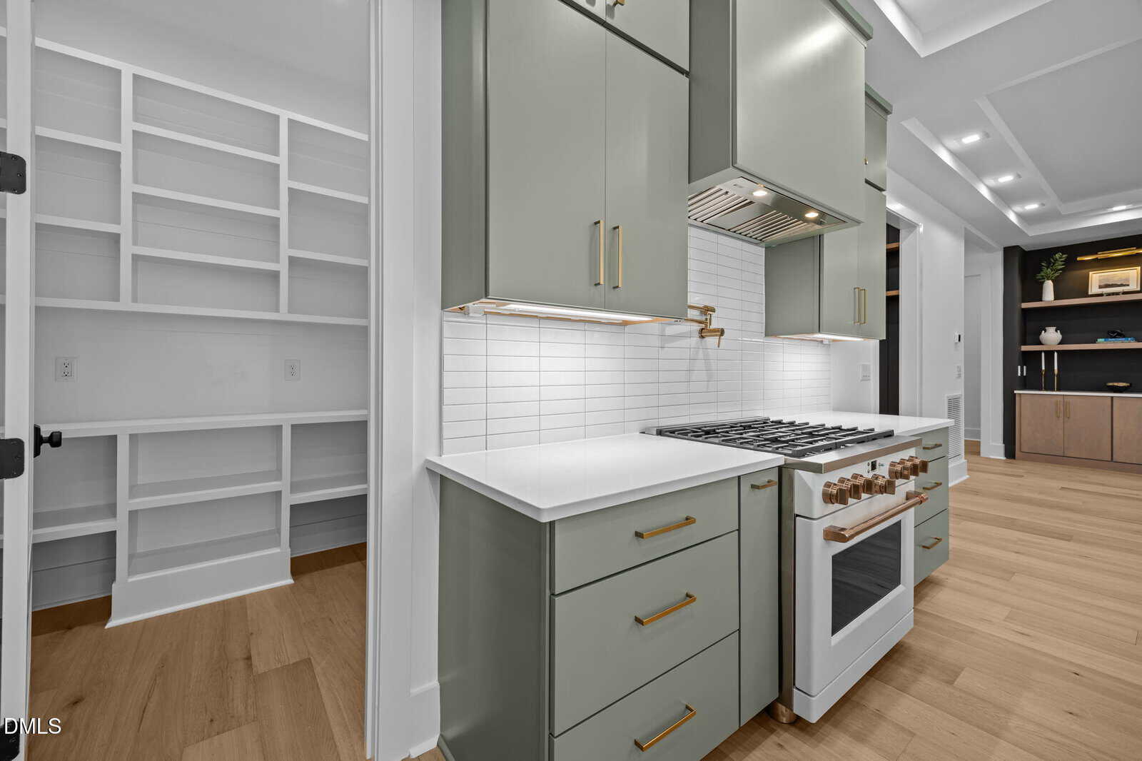 Modern kitchen with sage green cabinets, white countertops, gold hardware, a white tile backsplash, and a gas stove. An open pantry with built-in white shelves is visible to the left. Light wood flooring throughout.