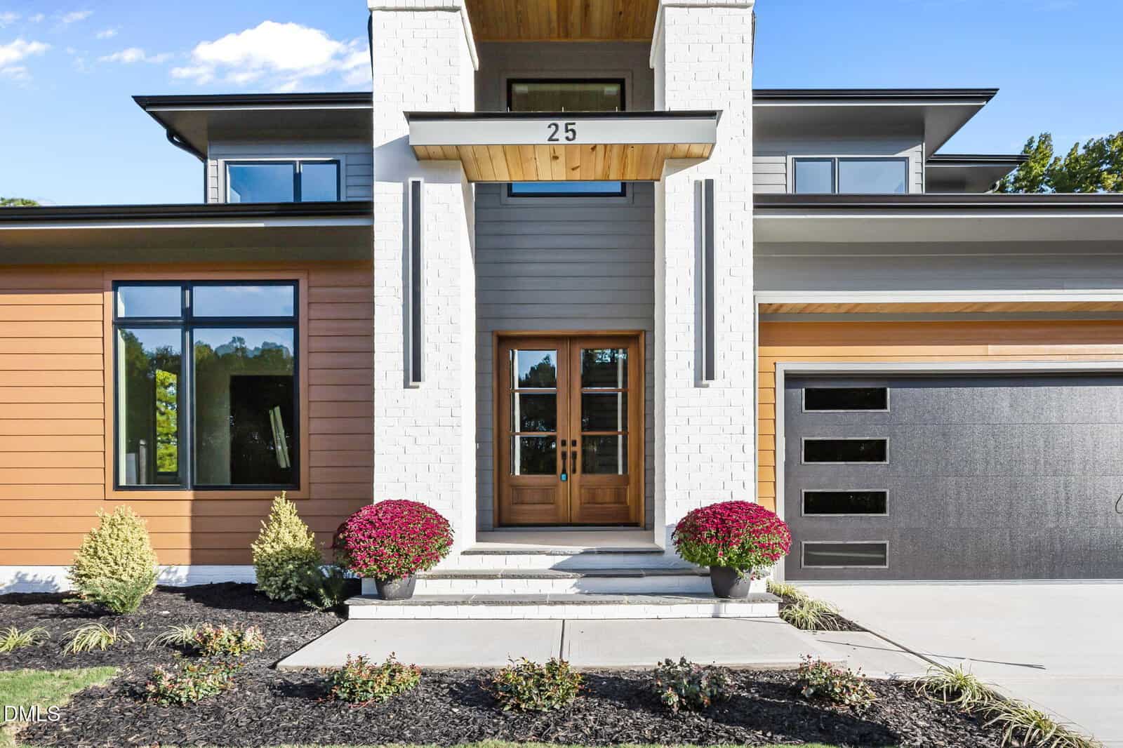 Modern house exterior with white brick and wood accents, large glass front door, black garage door, and two potted red mums by the entrance. Lush landscaping and clear blue sky in the background.