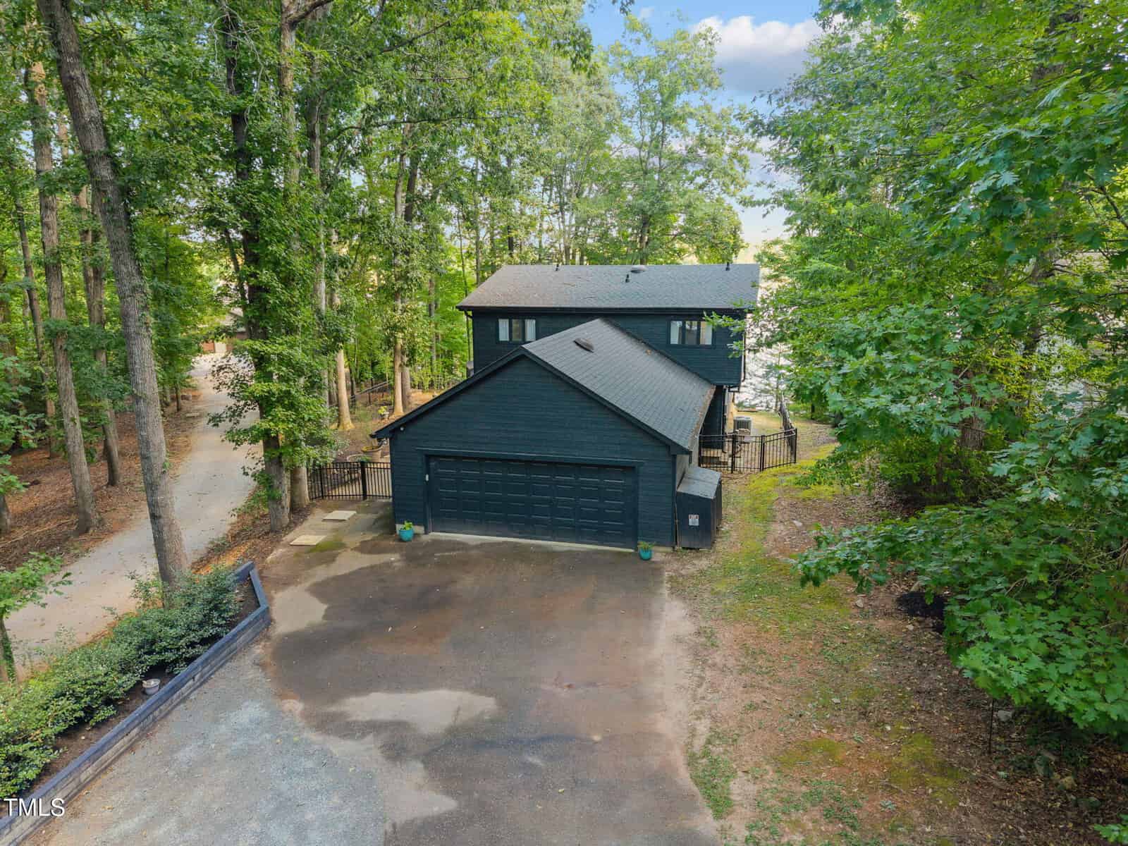 A two-story dark-colored house with a double garage sits surrounded by tall trees and greenery. A paved driveway leads to the house, and a quiet road runs alongside the property.