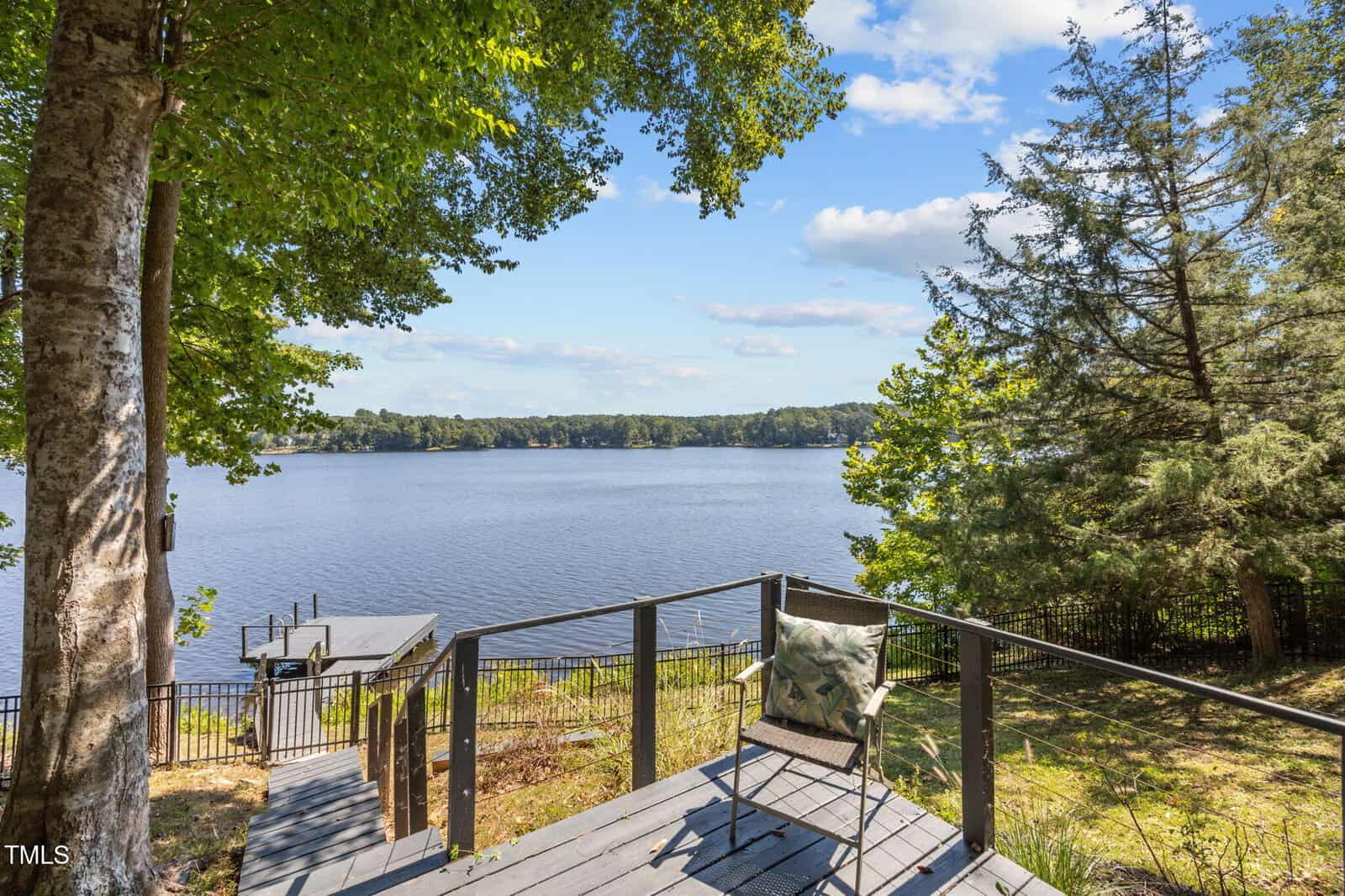 A peaceful lakeside scene with a deck featuring a chair and pillow overlooking a calm lake, surrounded by trees and greenery under a partly cloudy blue sky. A small dock extends into the water on the left.