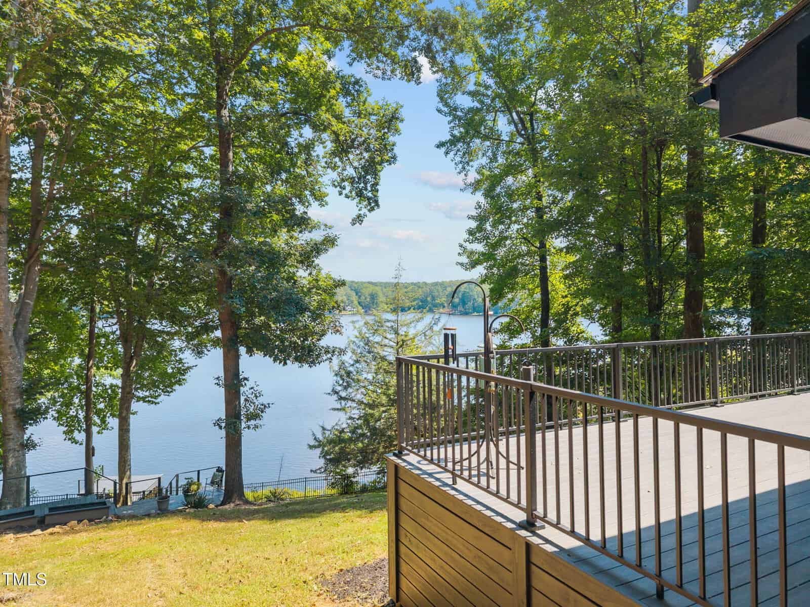 View from a wooden deck overlooking a peaceful lake surrounded by tall trees, with sunlight streaming through the leaves and a boat dock visible near the shoreline.