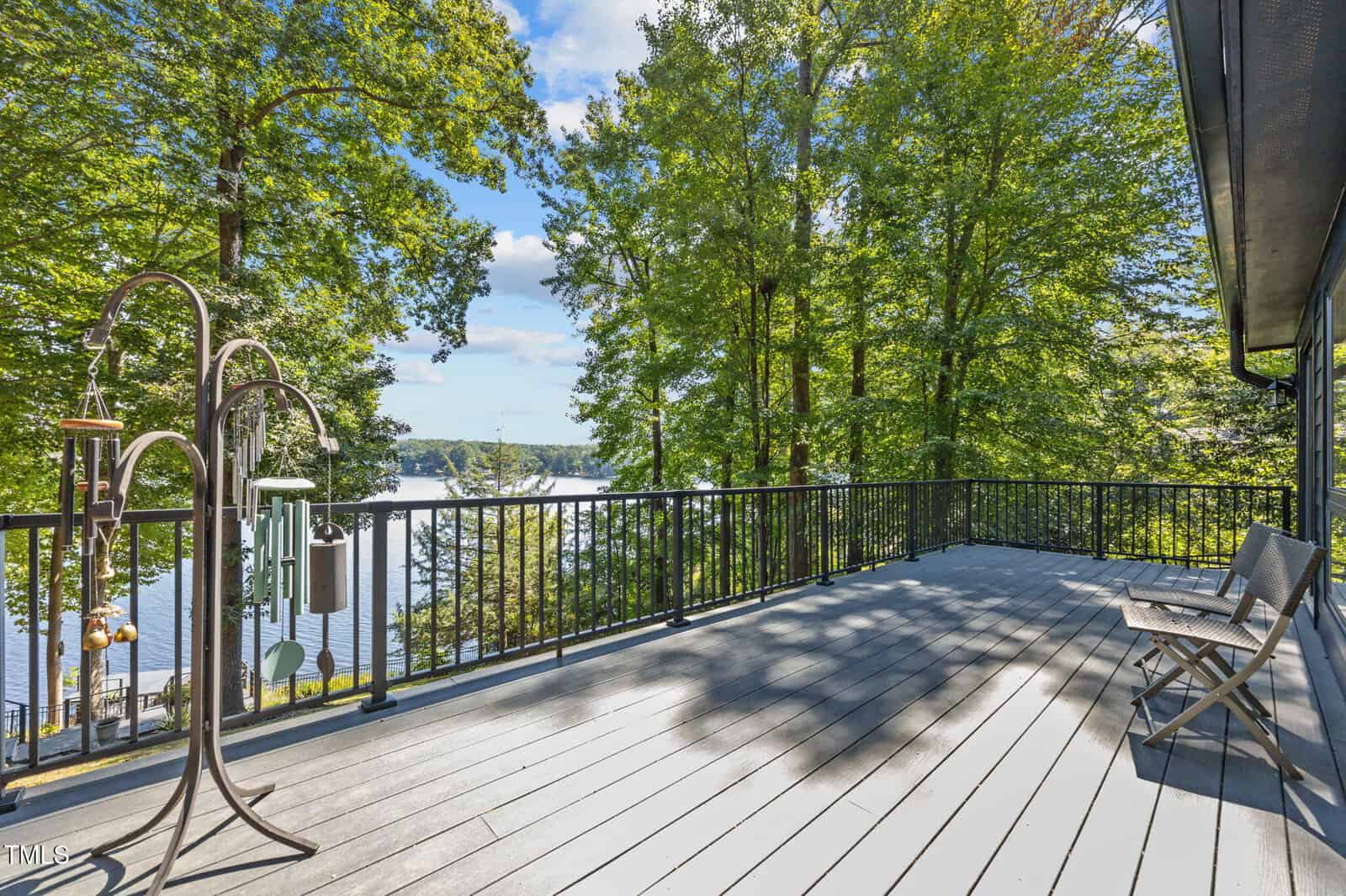 Spacious deck with metal railing, two wooden chairs, and wind chimes overlooks a scenic lake surrounded by tall green trees under a blue sky with scattered clouds.