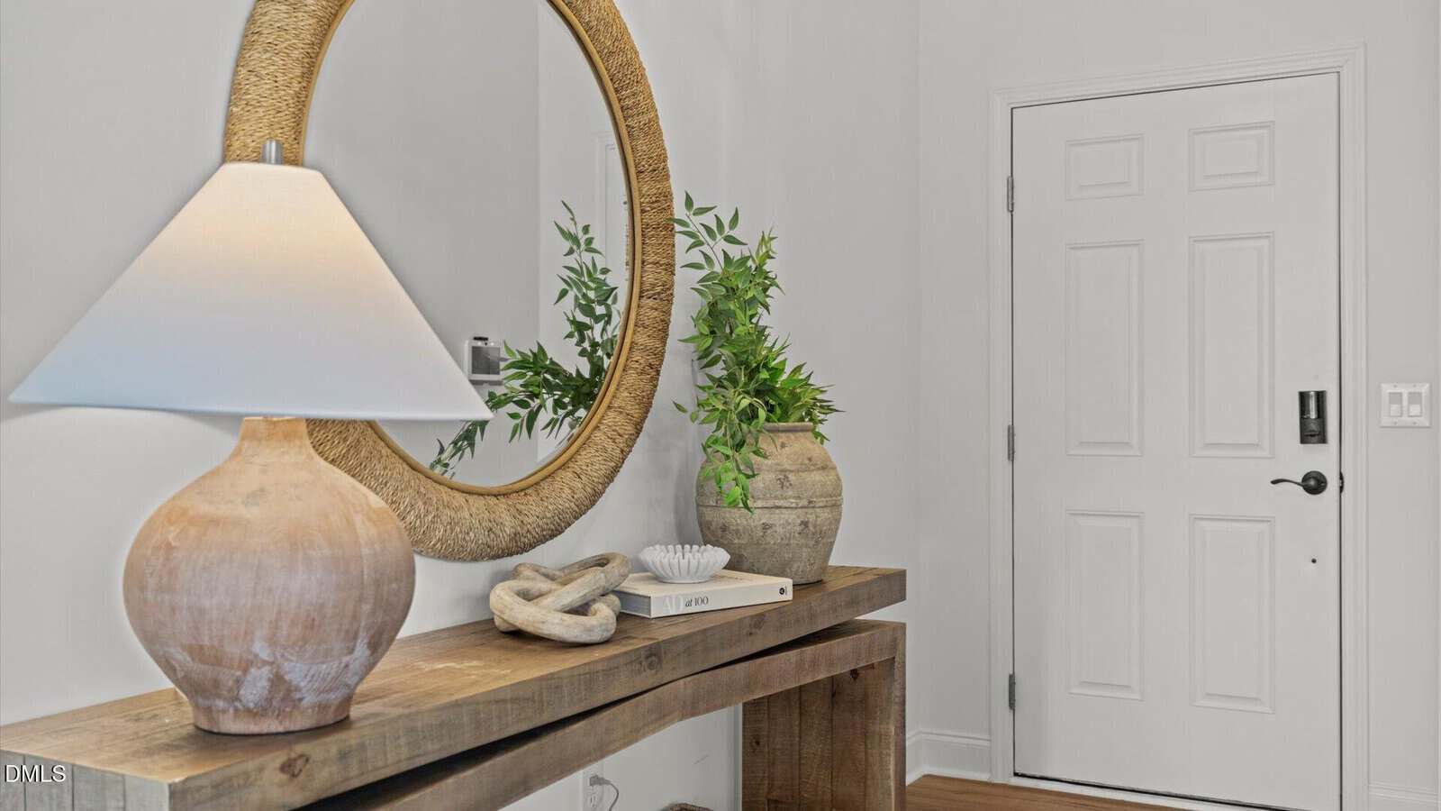 A wooden console table with a ceramic lamp, decorative objects, and a plant sits below a round mirror near a white front door in a bright entryway.