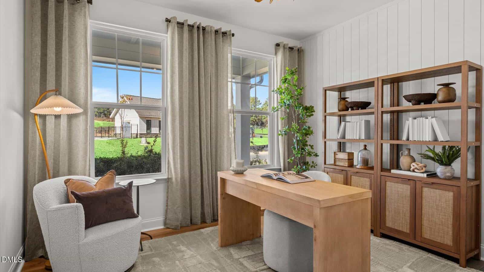 A home office with a wooden desk, light gray chair, bookshelves filled with books and decor, tall curtains, a modern floor lamp, a potted plant, and large windows showing a house and greenery outside.