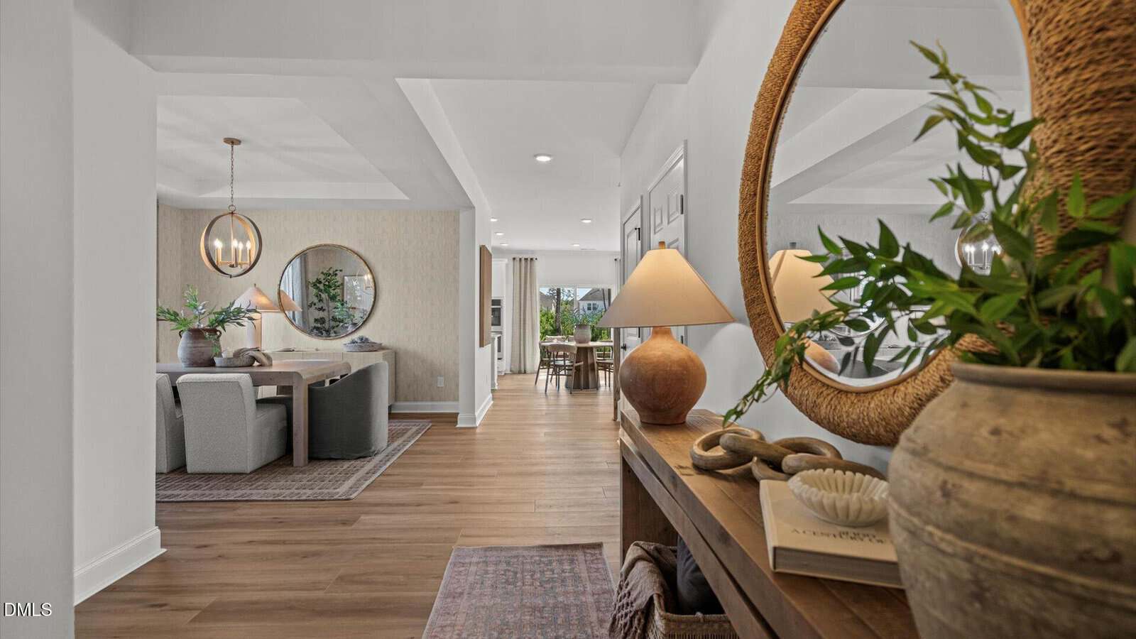 A modern home interior with light wood floors, a hallway console table with decor and a large round mirror, and a dining area with a round table, upholstered chairs, and another round mirror on the wall.