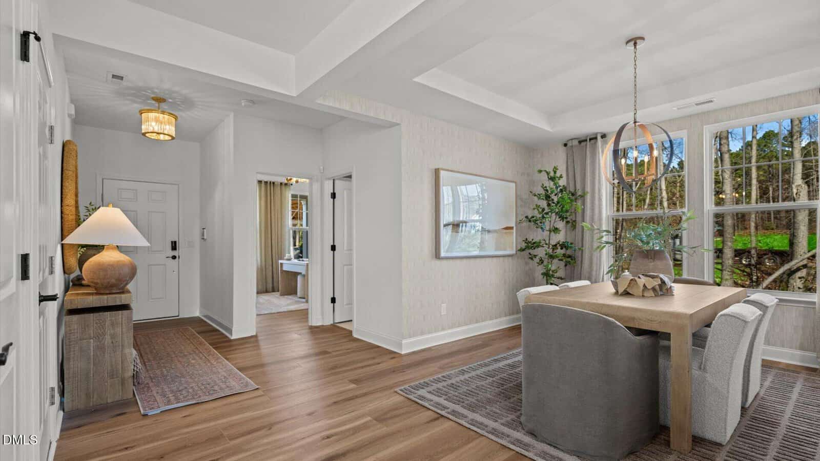 A dining area with a wooden table and six chairs sits next to large windows. Adjacent is a hallway with a white front door, overhead lighting, a rug, and a glimpse of a sink through an open door.