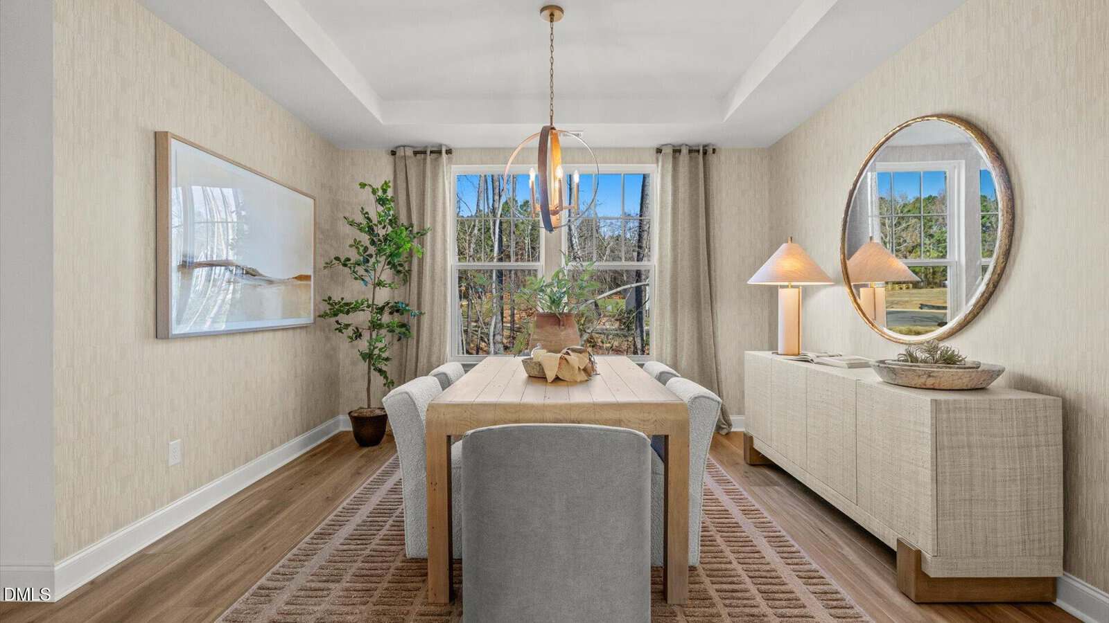 Modern dining room with a wooden table, six chairs, a sideboard with two lamps, round mirror, large plant, wall art, and windows showing trees outside. Neutral tones and natural light create a cozy, inviting space.