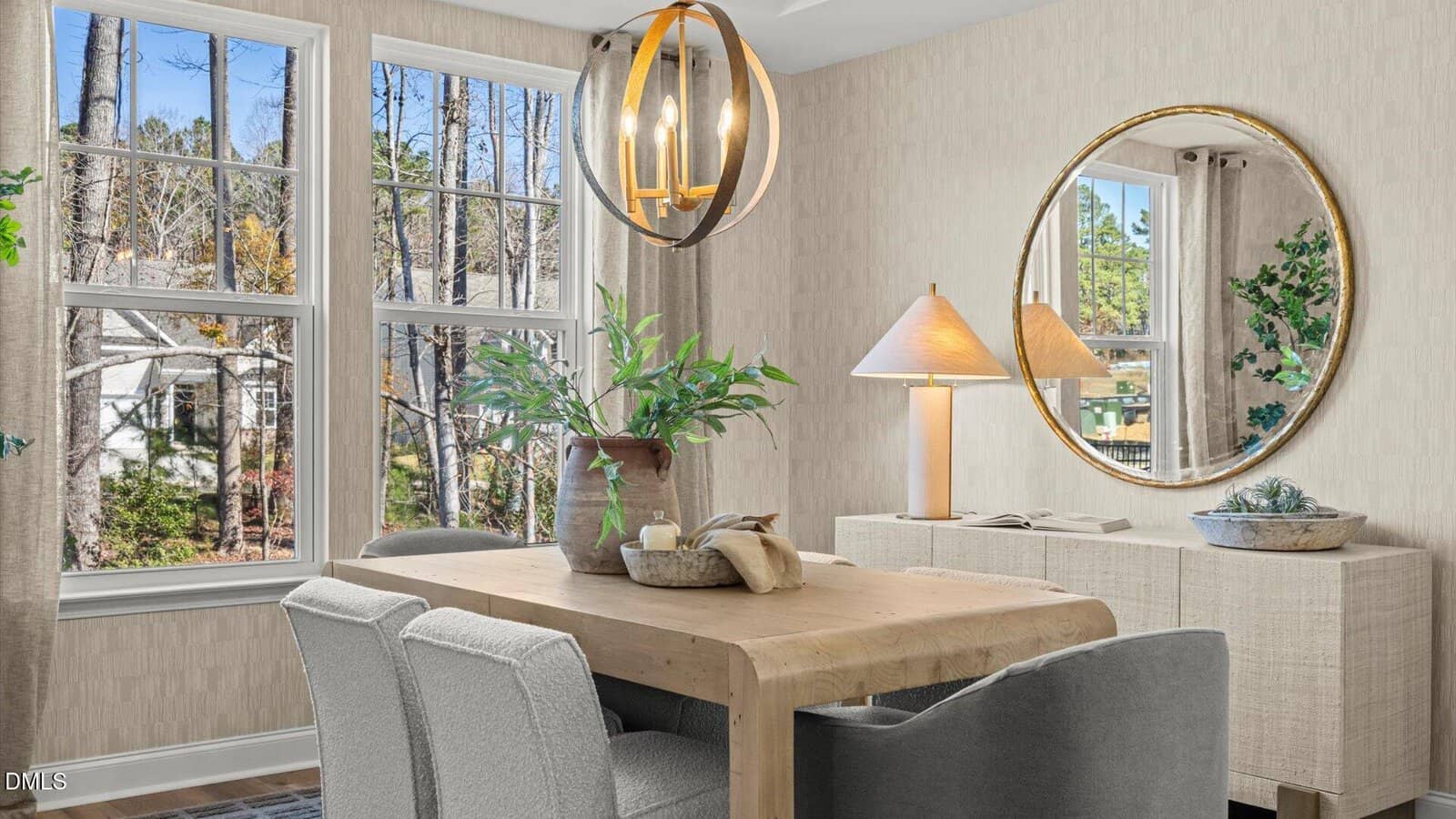 Bright dining room with a wooden table, gray chairs, modern chandelier, large round mirror, table lamp, and window view of trees and houses outside. Neutral tones create a cozy, inviting atmosphere.